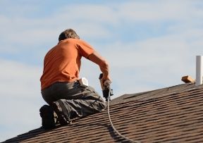 Roofer kneeling on roof, using a nail gun; blue sky in background.