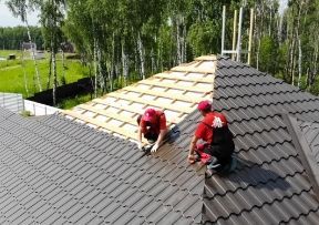 Two workers installing a dark metal roof on a house, one side complete.
