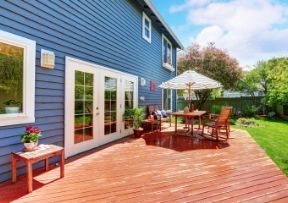 Blue house with wooden deck, table, chairs, and umbrella in sunny backyard.