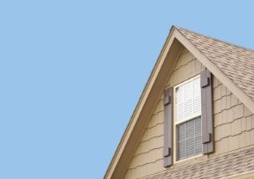 Tan house gable with window and brown shutters, against a blue sky.