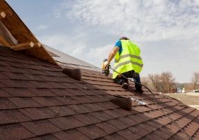Roofer in safety vest using a nail gun on a brown shingled roof, outdoors on a sunny day.