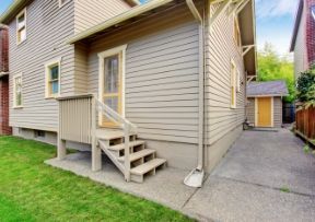 Beige house exterior with steps leading to a door, a shed in the background, and a concrete driveway.