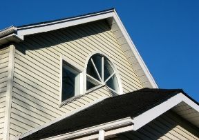 Beige-sided house with a dark roof and a triangular window against a clear blue sky.