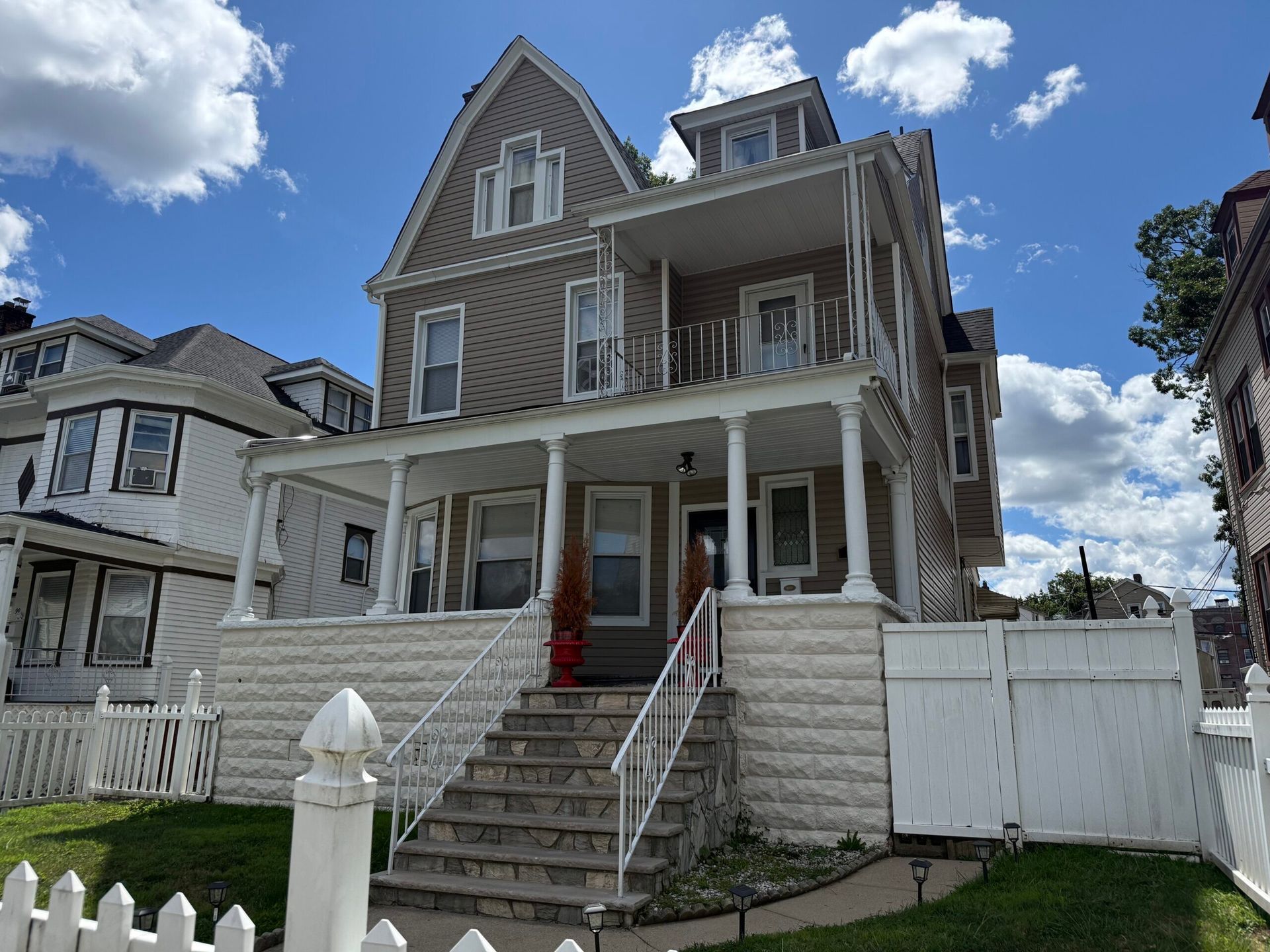 Brown and white Victorian house with a two-story porch, white picket fence, and blue sky.