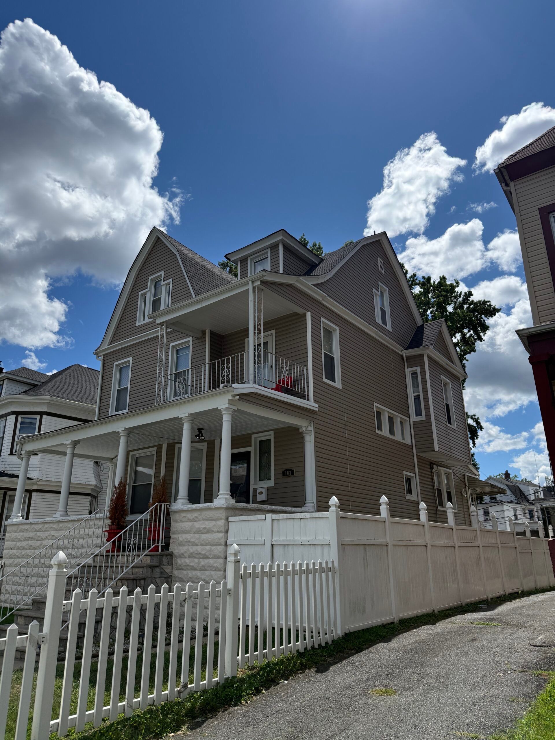 Tan multi-story house with white trim, porch, and fence against a blue sky with fluffy clouds.