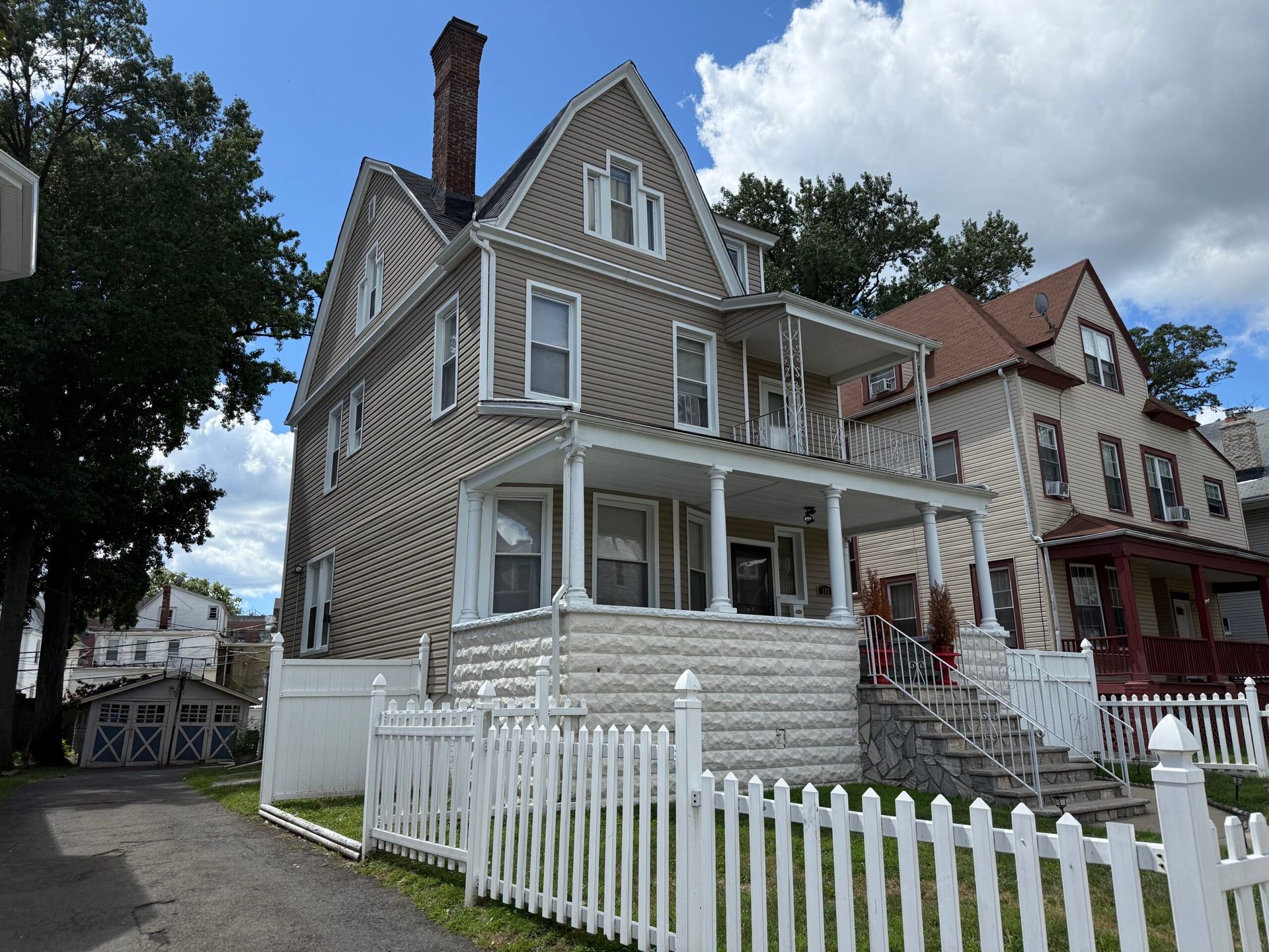 Three-story beige house with white picket fence, porch, and chimney. Another house is visible to the right.