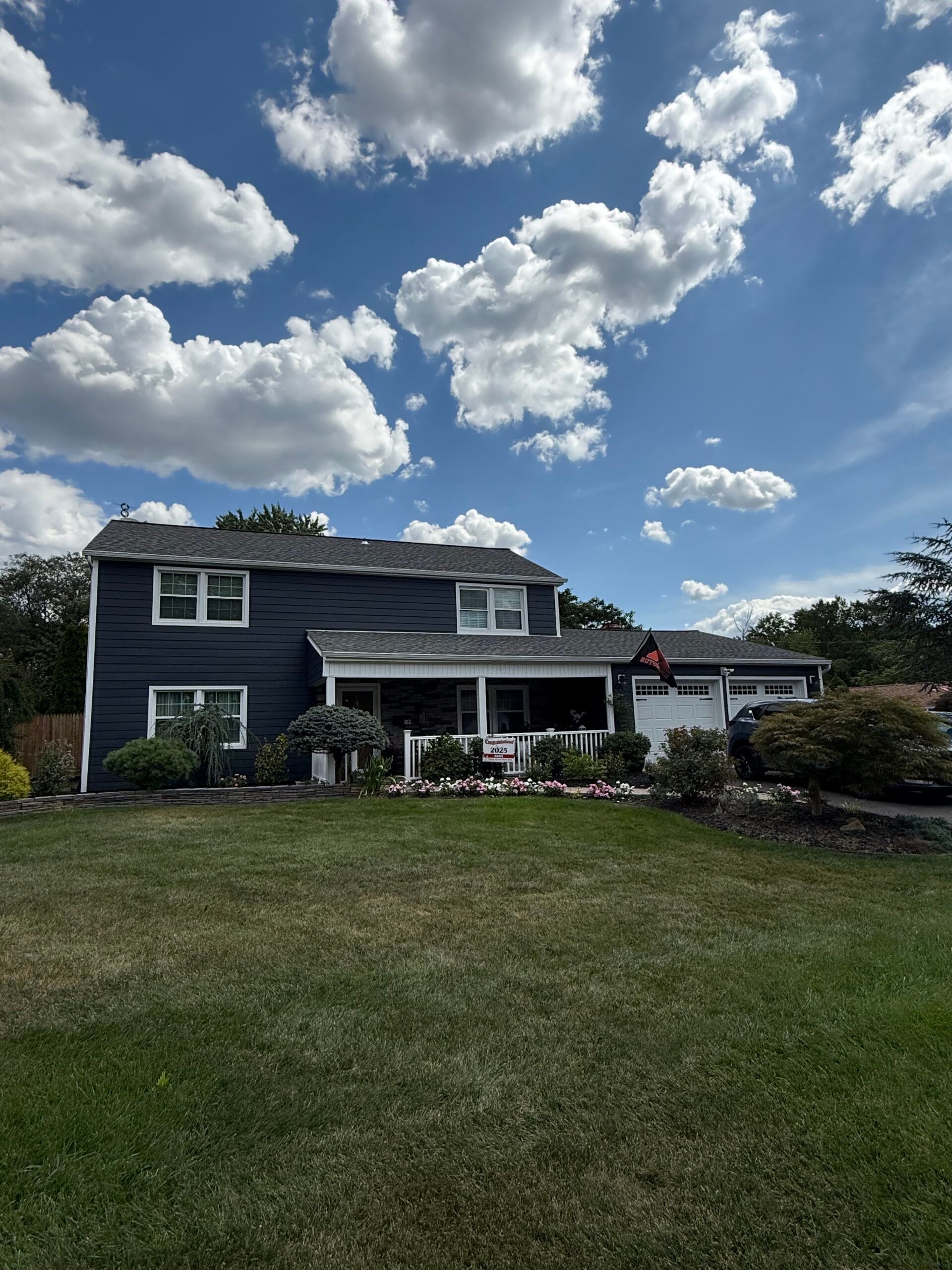 Two-story blue house with white trim under a partly cloudy sky. Green lawn and shrubs in front.