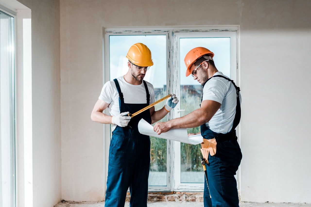 Two construction workers in a room reviewing blueprints, one with a tape measure.