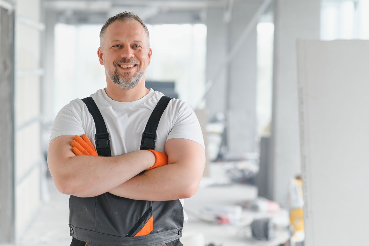 Construction worker in white shirt and overalls, arms crossed, smiling in a construction site.