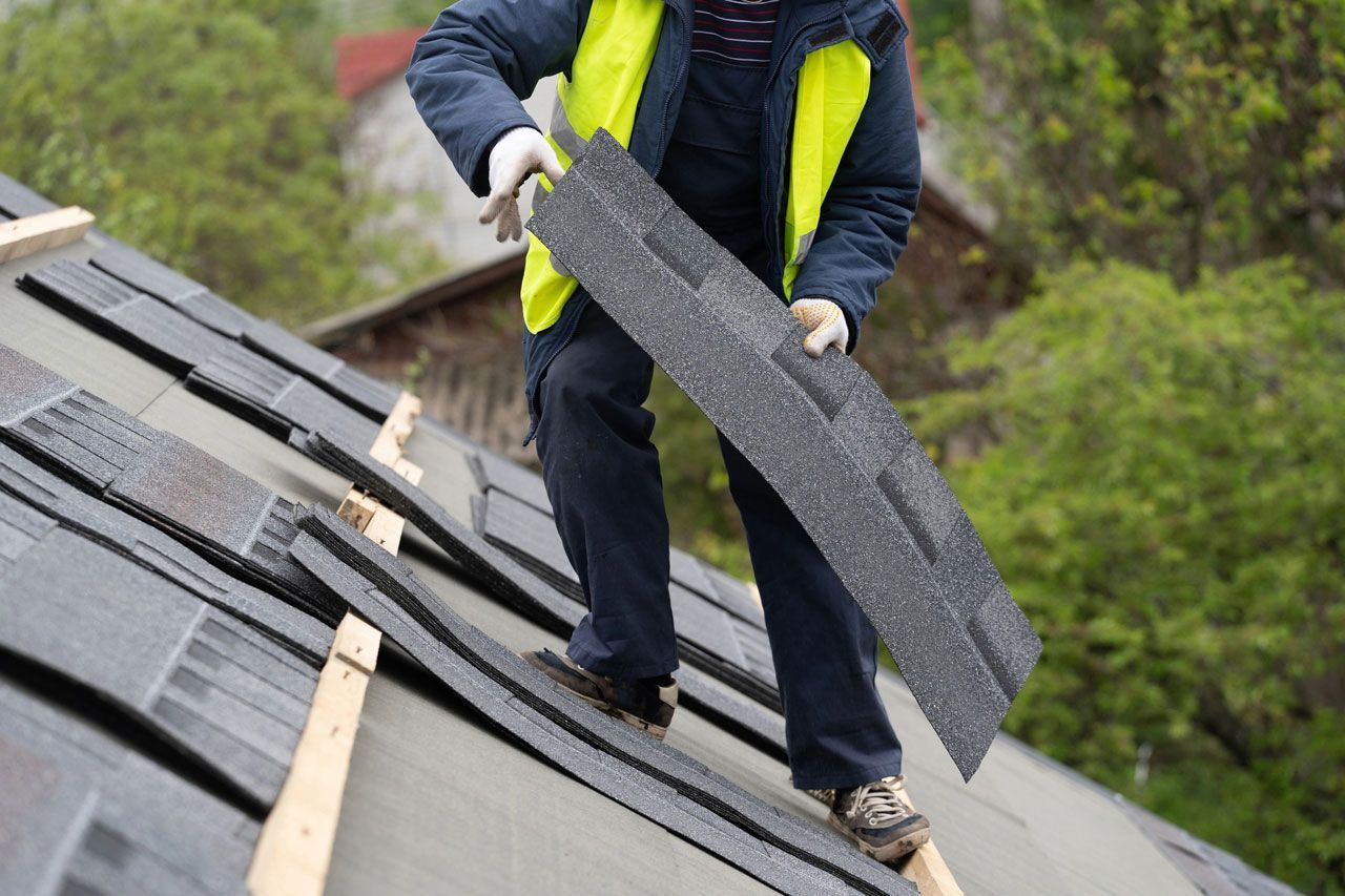 Roofer on a roof, installing asphalt shingles, wearing work clothes and a yellow safety vest.