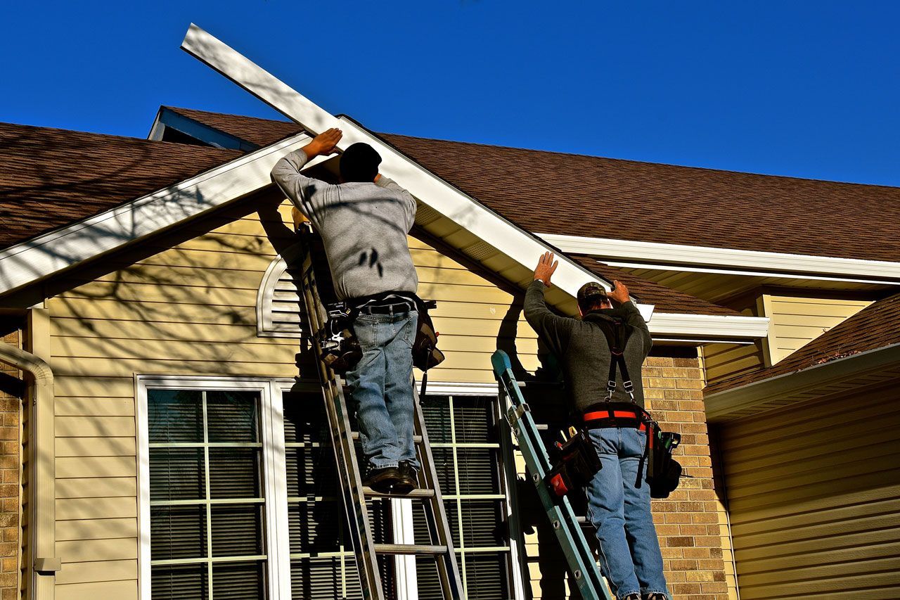 Two workers installing a white gutter on a house with brown roof and siding; blue sky.