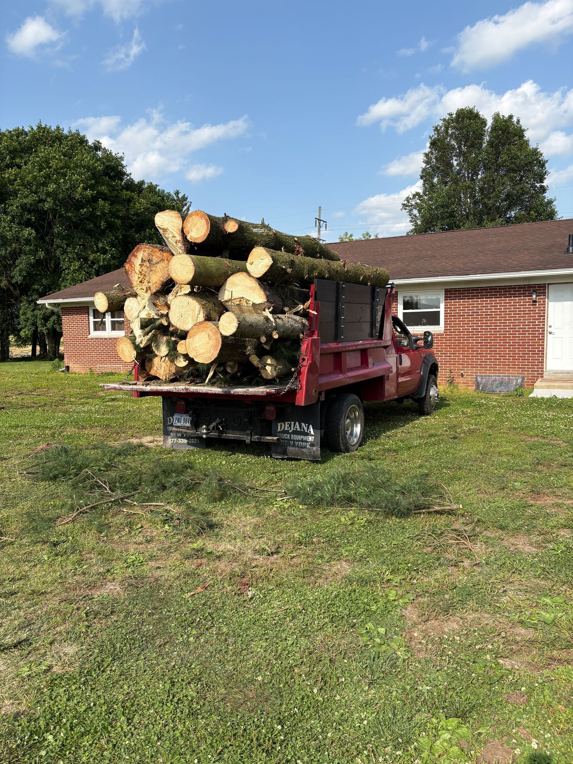 Red pickup truck loaded with logs in front of a house on a grassy lawn under a blue sky.