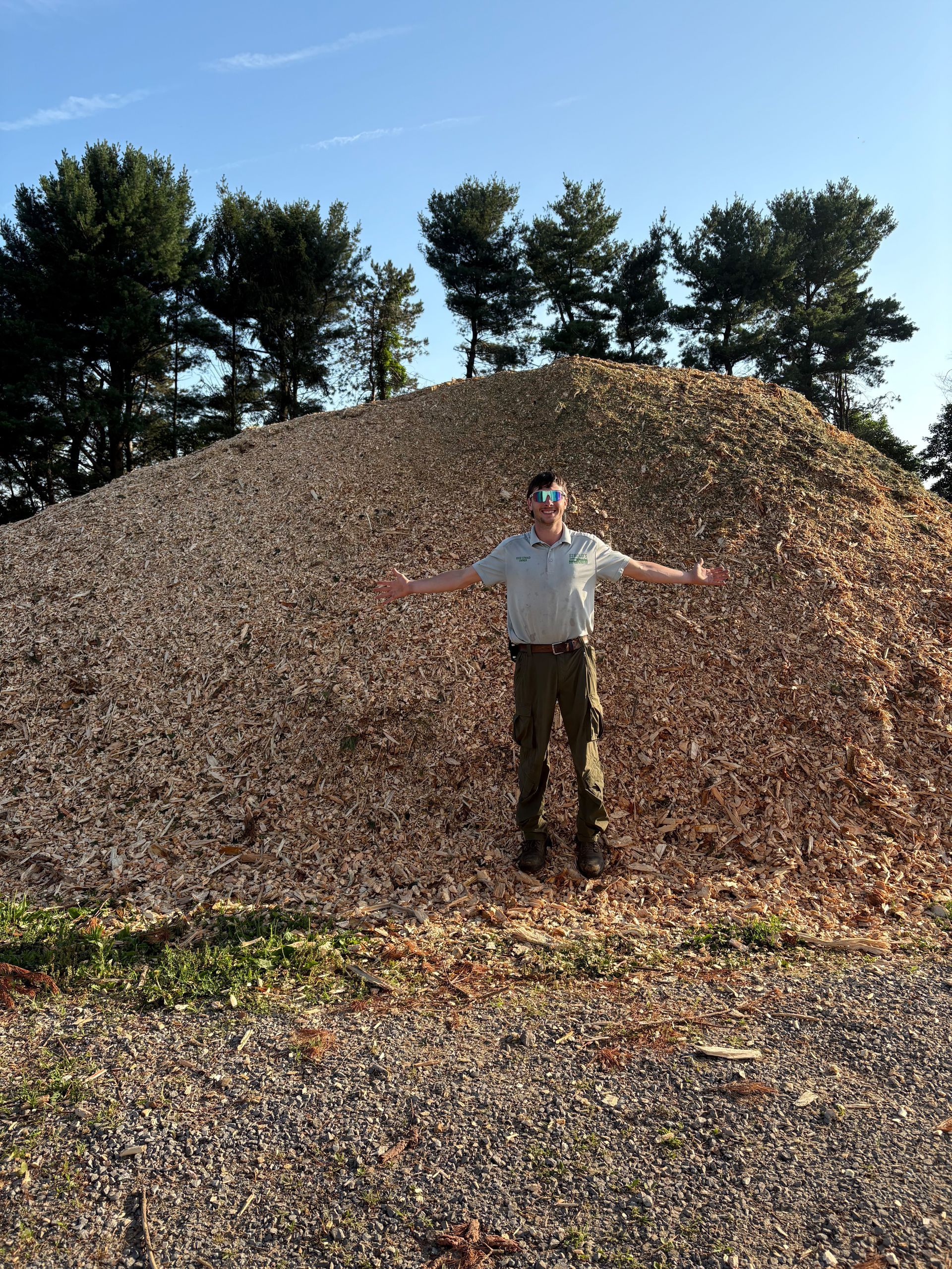 Man standing in front of a large pile of wood chips with arms outstretched, blue sky in background.