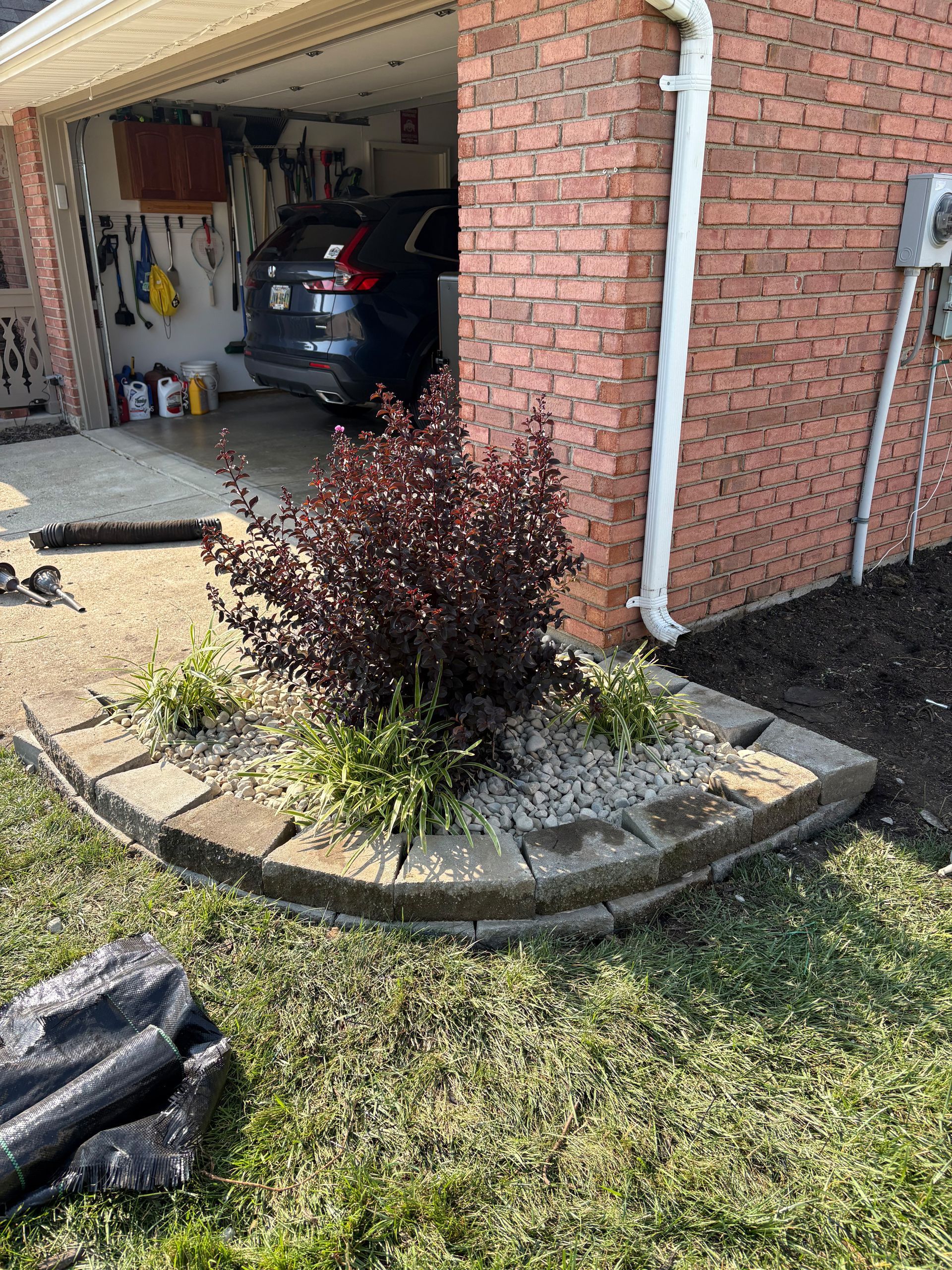A landscaping bed with a red bush, gray pavers, and a brick wall next to a garage.