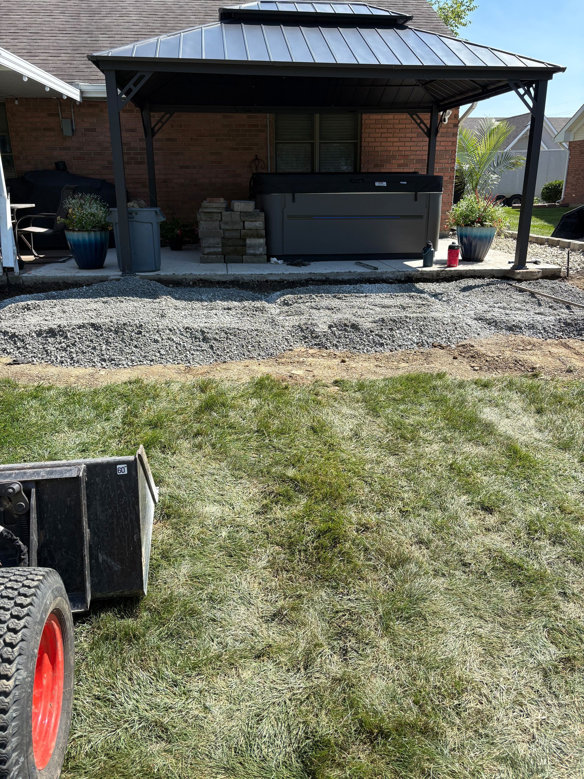A hot tub under a gazebo, surrounded by gravel and grass. A tractor is in the foreground.