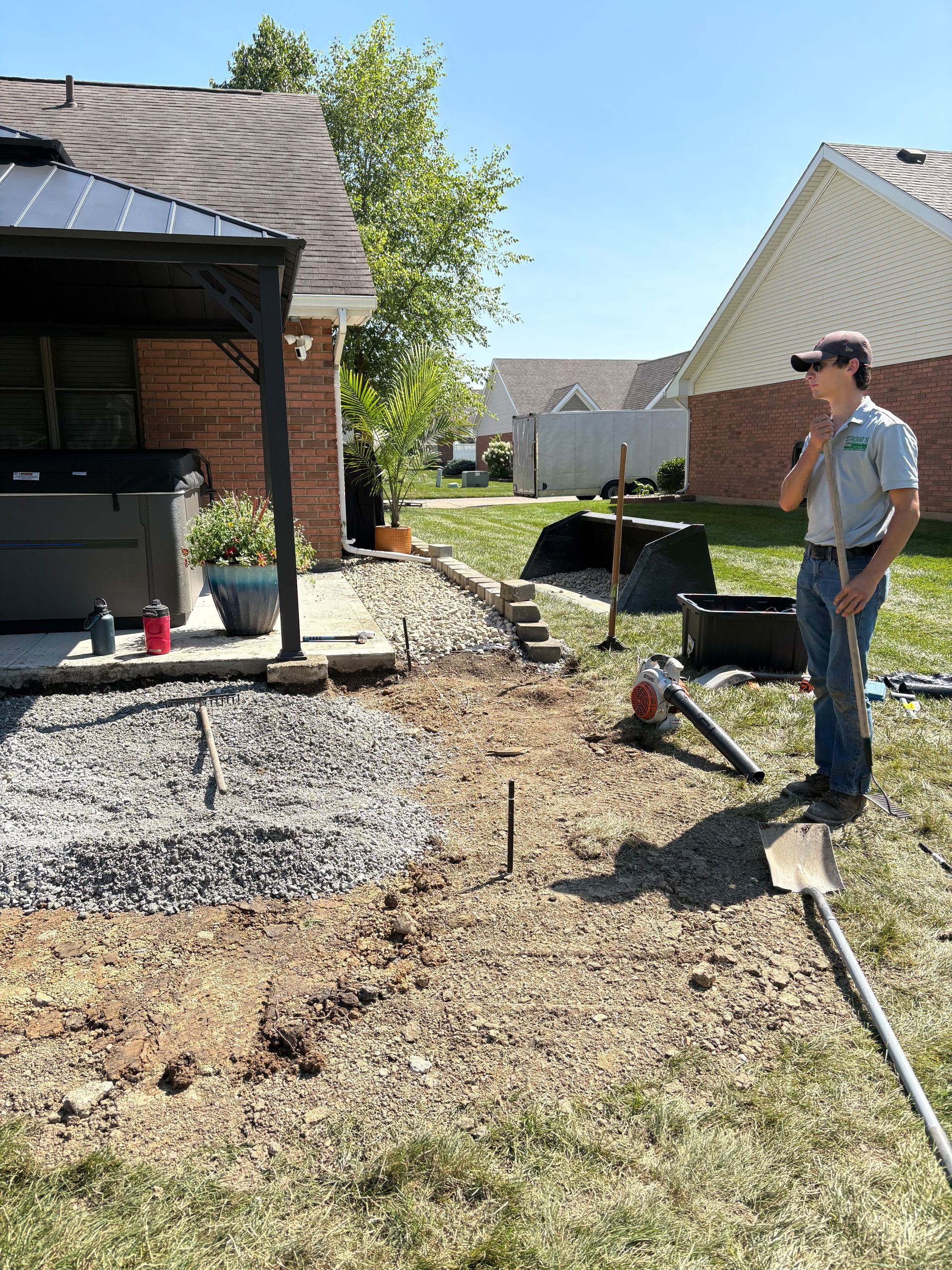 Man in a yard with tools, landscaping near a house with a hot tub and gazebo.