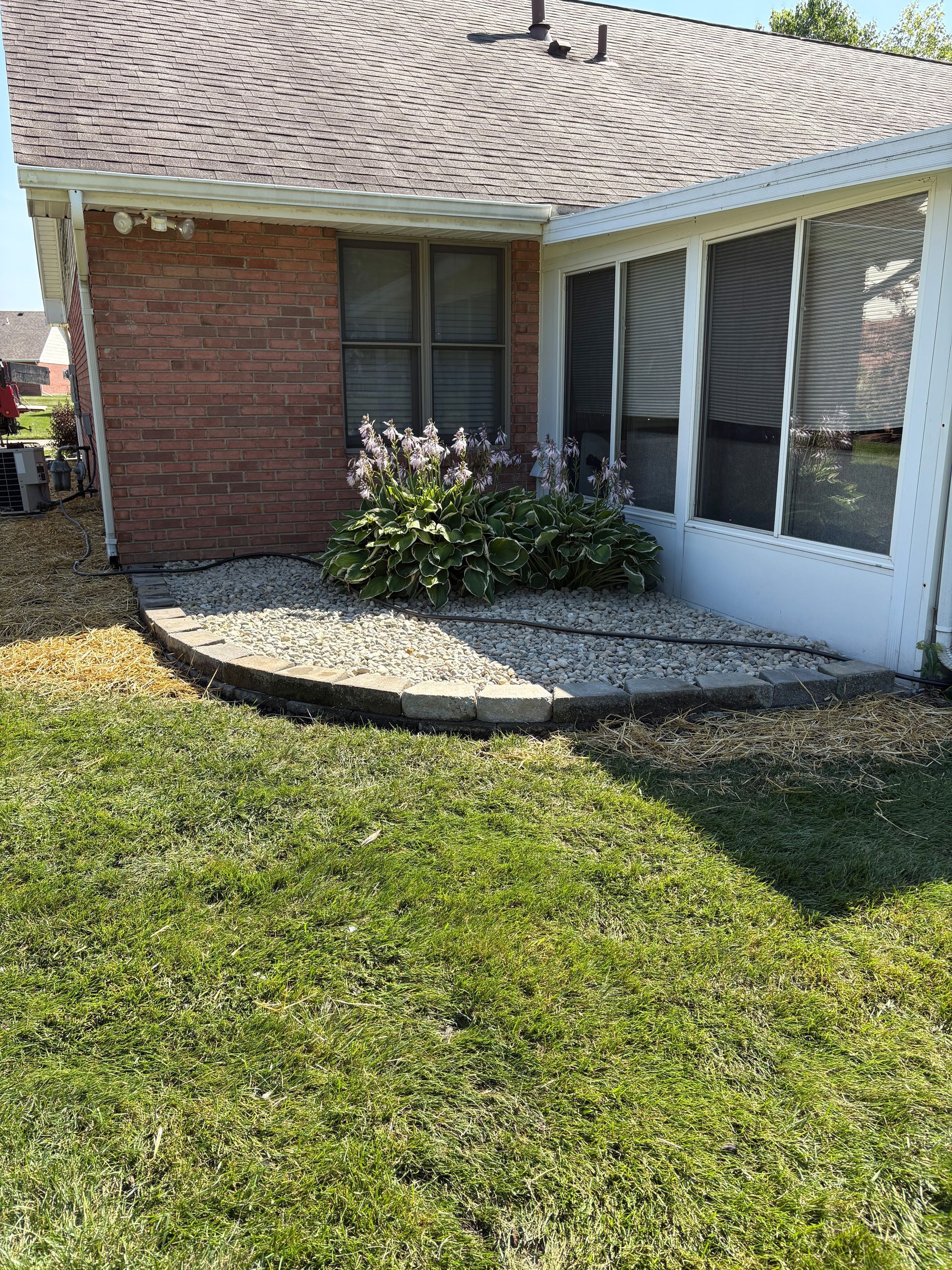 Backyard with brick wall, screened porch, and stone-bordered flower bed with green plants. Green grass in foreground.