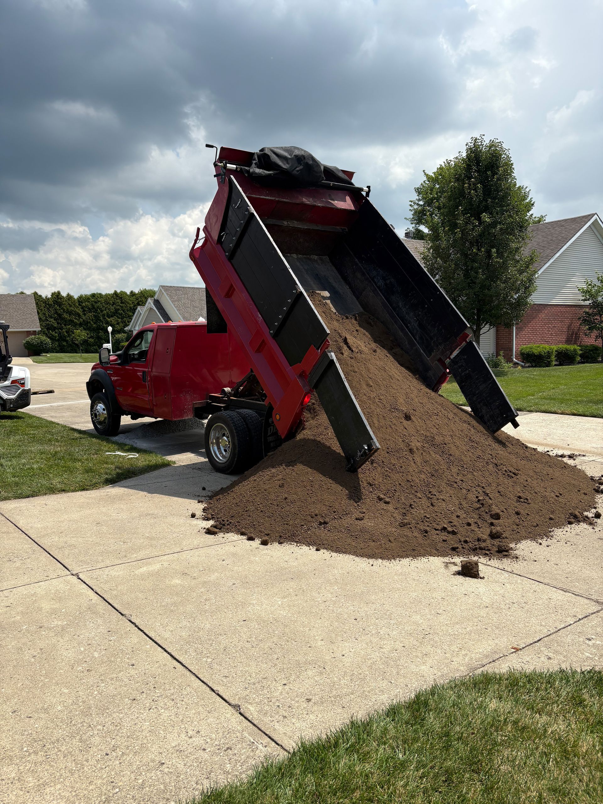 Red dump truck unloading dirt onto a driveway in a suburban neighborhood.