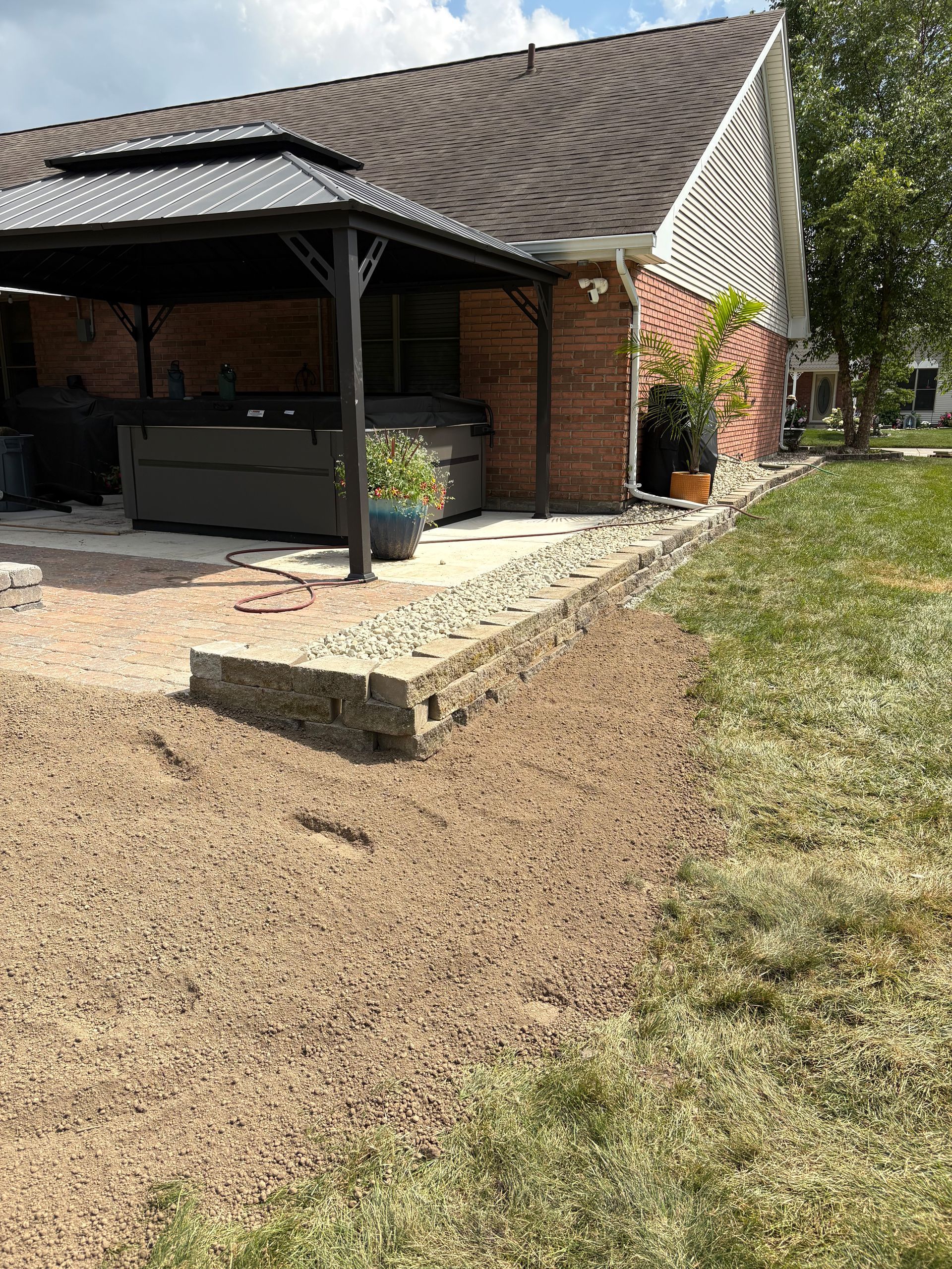 Patio with gazebo, hot tub, and gravel border next to a brick house with grass on the right.