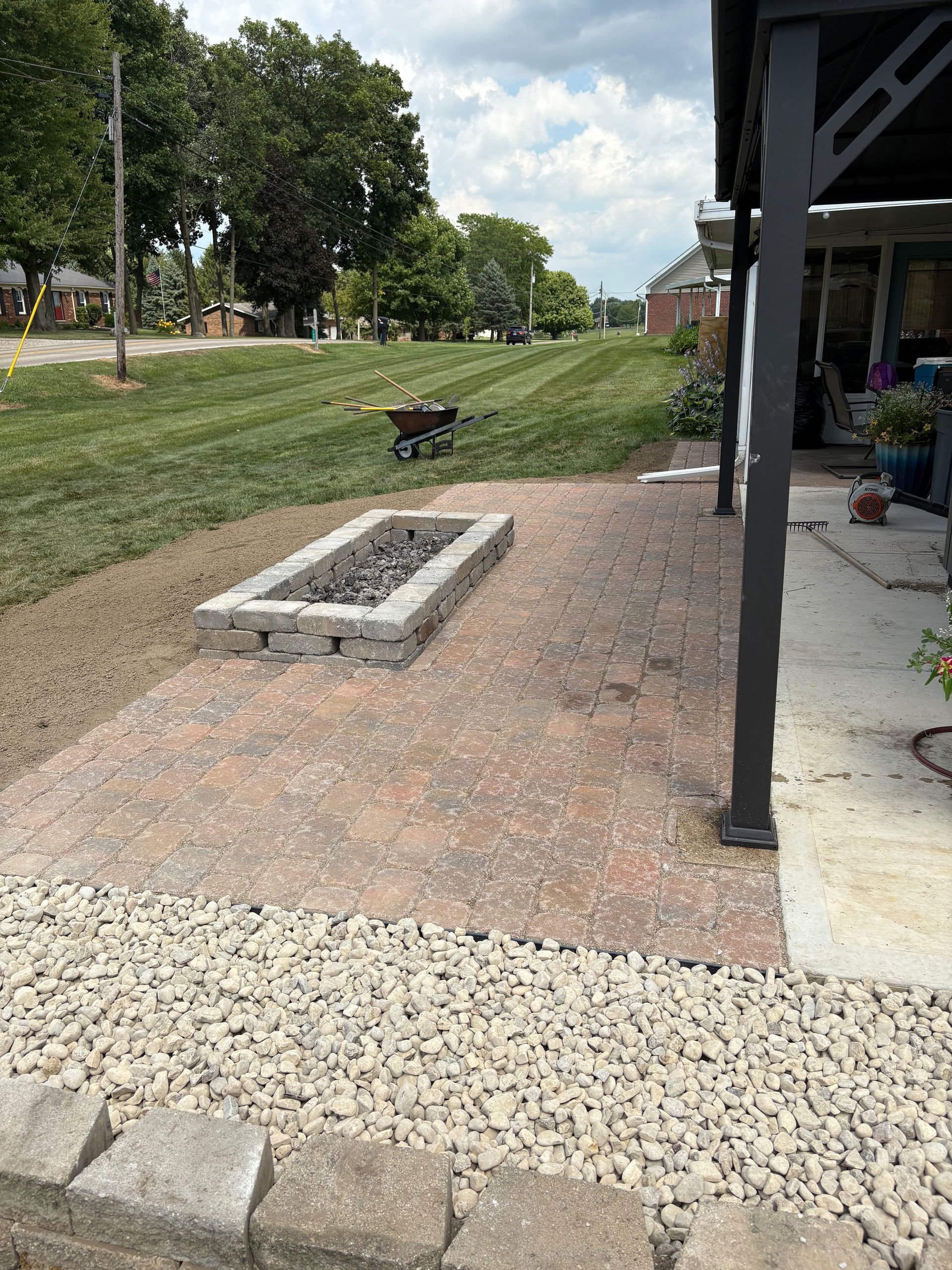 A backyard patio with a fire pit, bordered by gravel and low walls, next to a house and lawn.