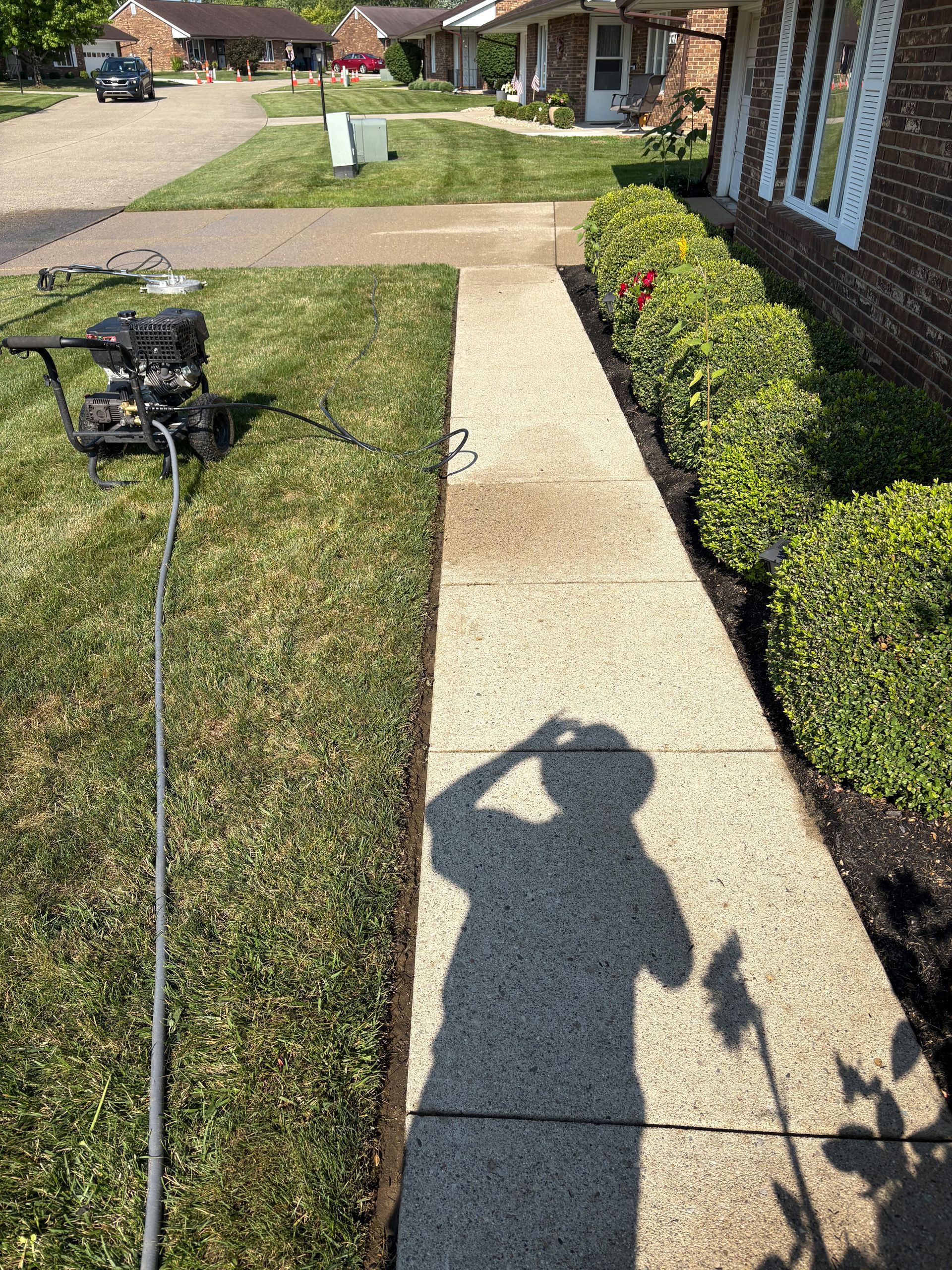 Person’s shadow on a concrete path alongside a lawn, with a mower and bushes; sunny outdoors.