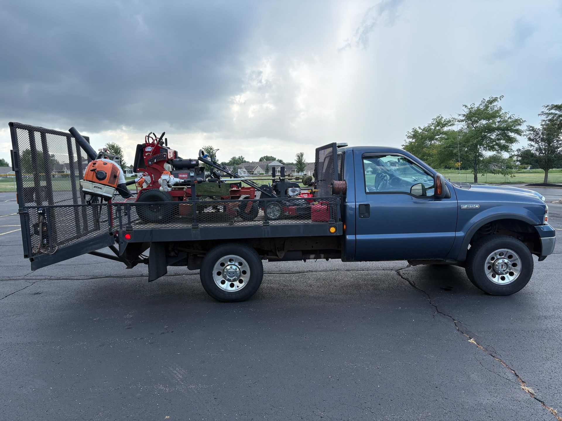 Blue pickup truck with landscaping equipment loaded on the bed, parked on pavement under a cloudy sky.