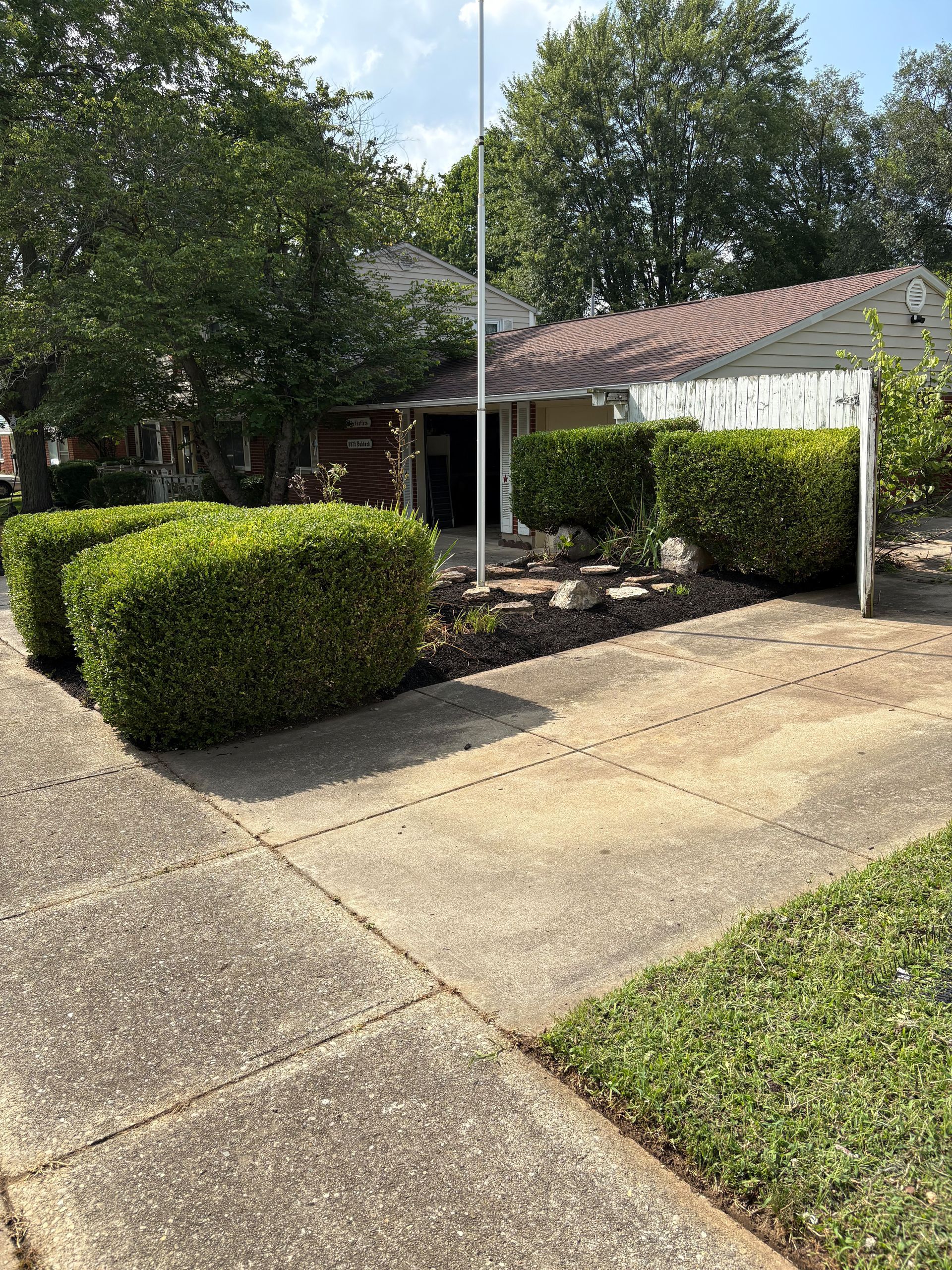 A house with a concrete driveway, green bushes, and a flagpole in front.