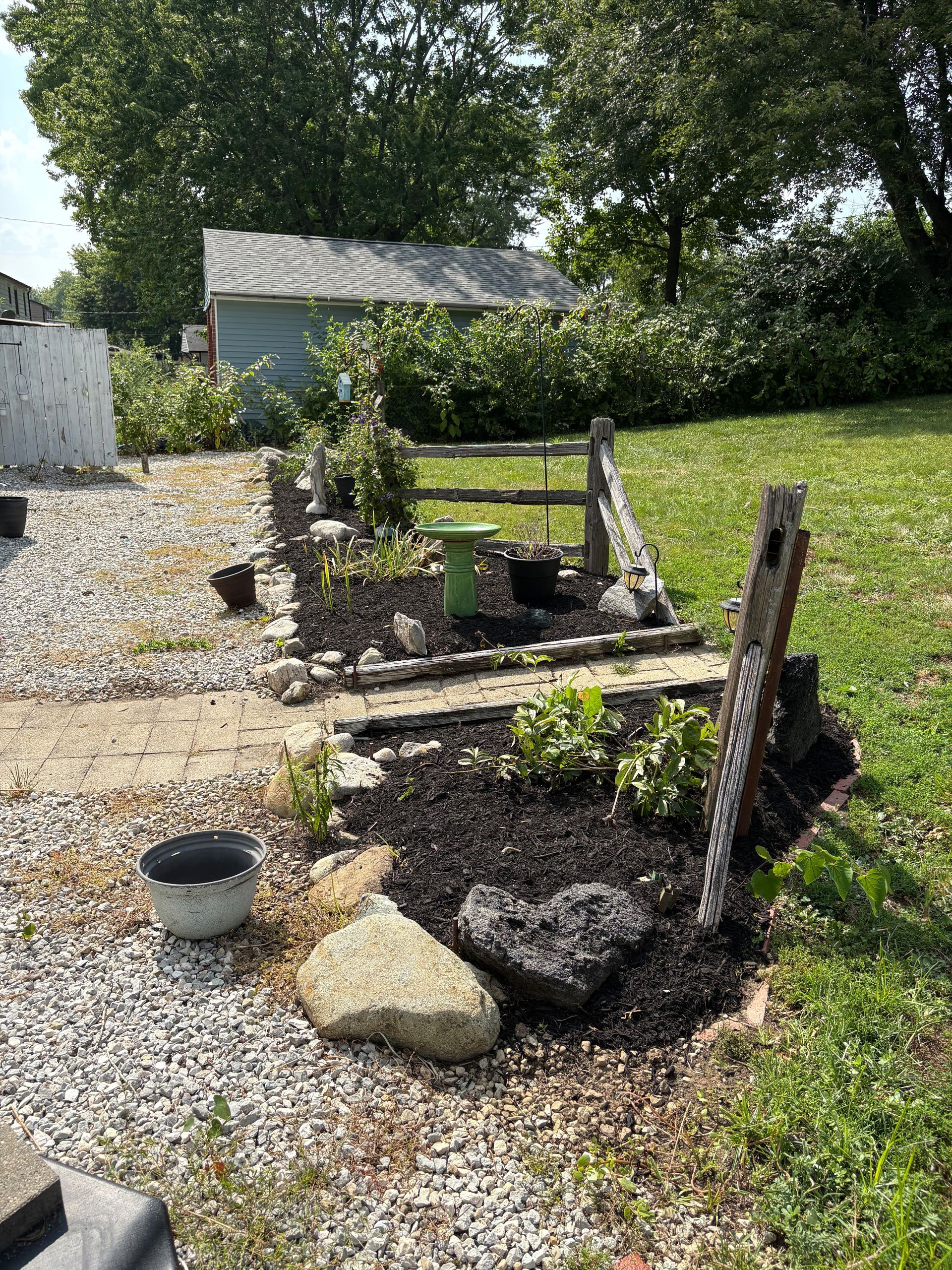 Backyard garden with flowers, mulch, and rocks; a weathered wooden fence and small shed in the background.