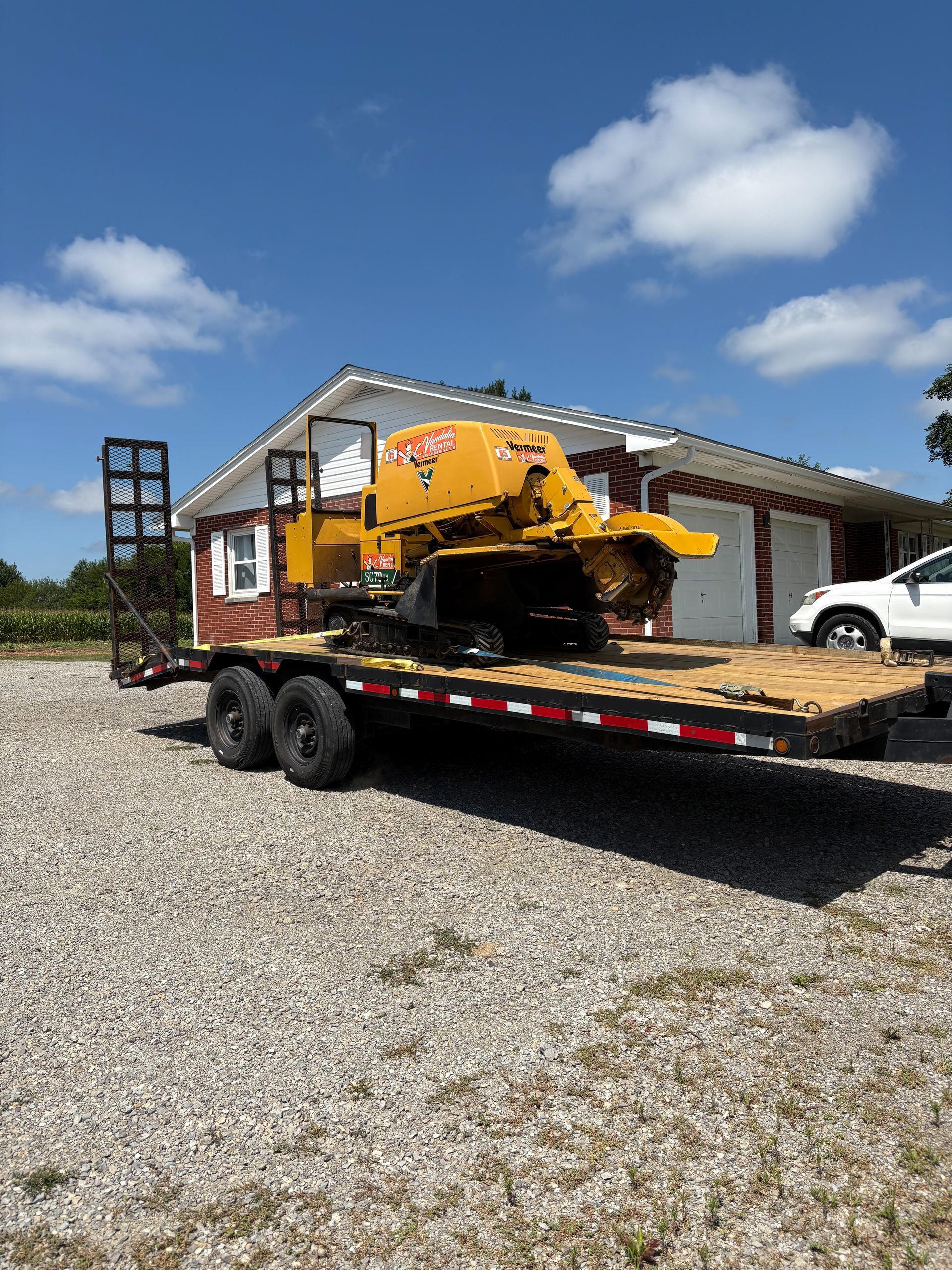 Yellow stump grinder on a flatbed trailer, parked in front of a house on a gravel lot under a blue sky.