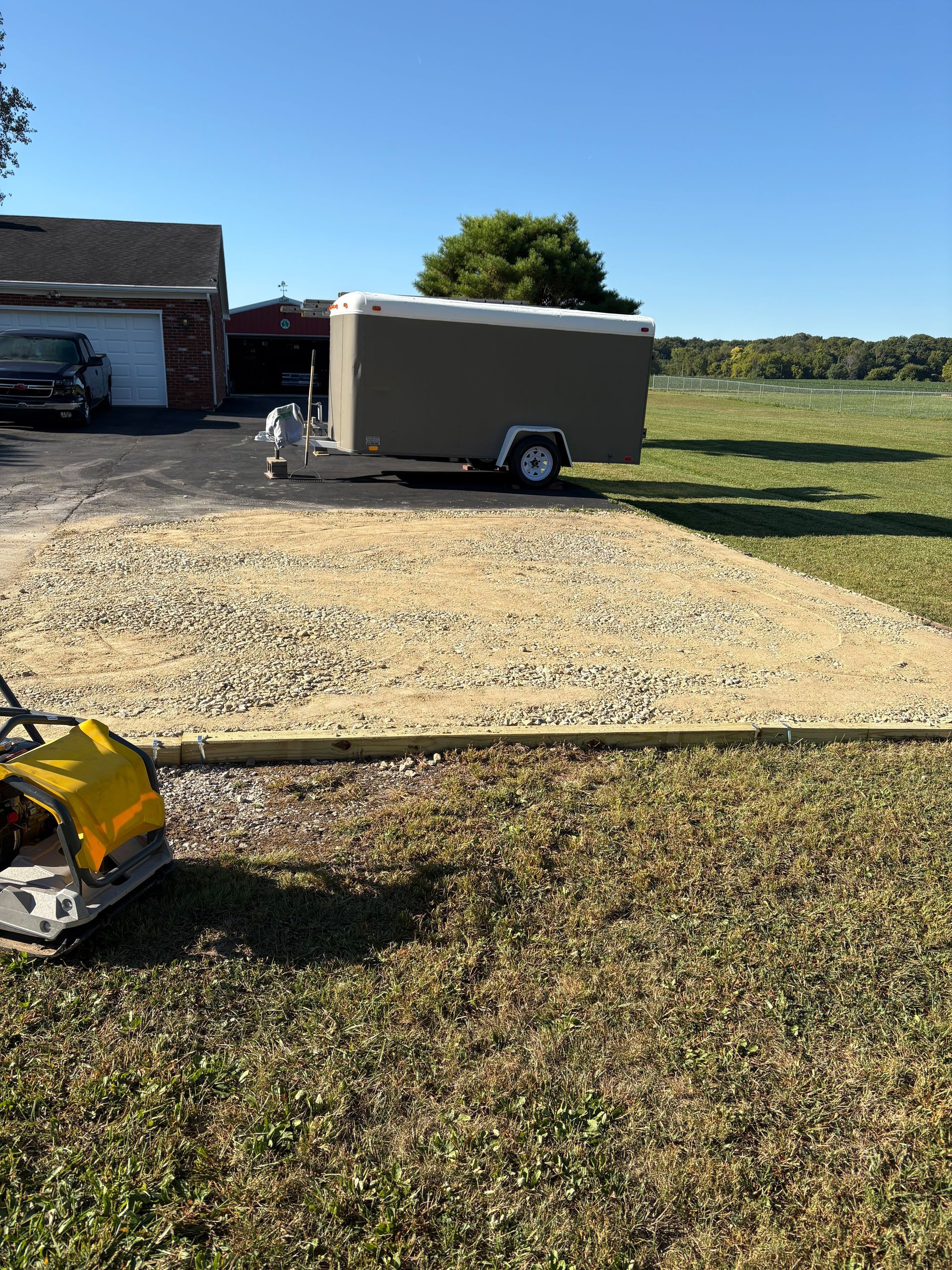 Gravel driveway under construction with a trailer parked on it, a compactor, and a house in the background.