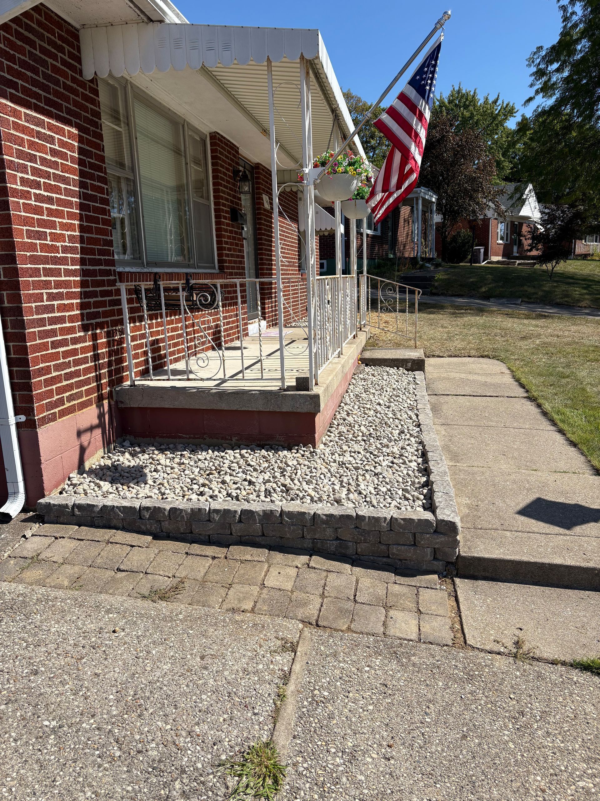 Brick house with porch, sidewalk, and a rock bed in front. American flag waving.