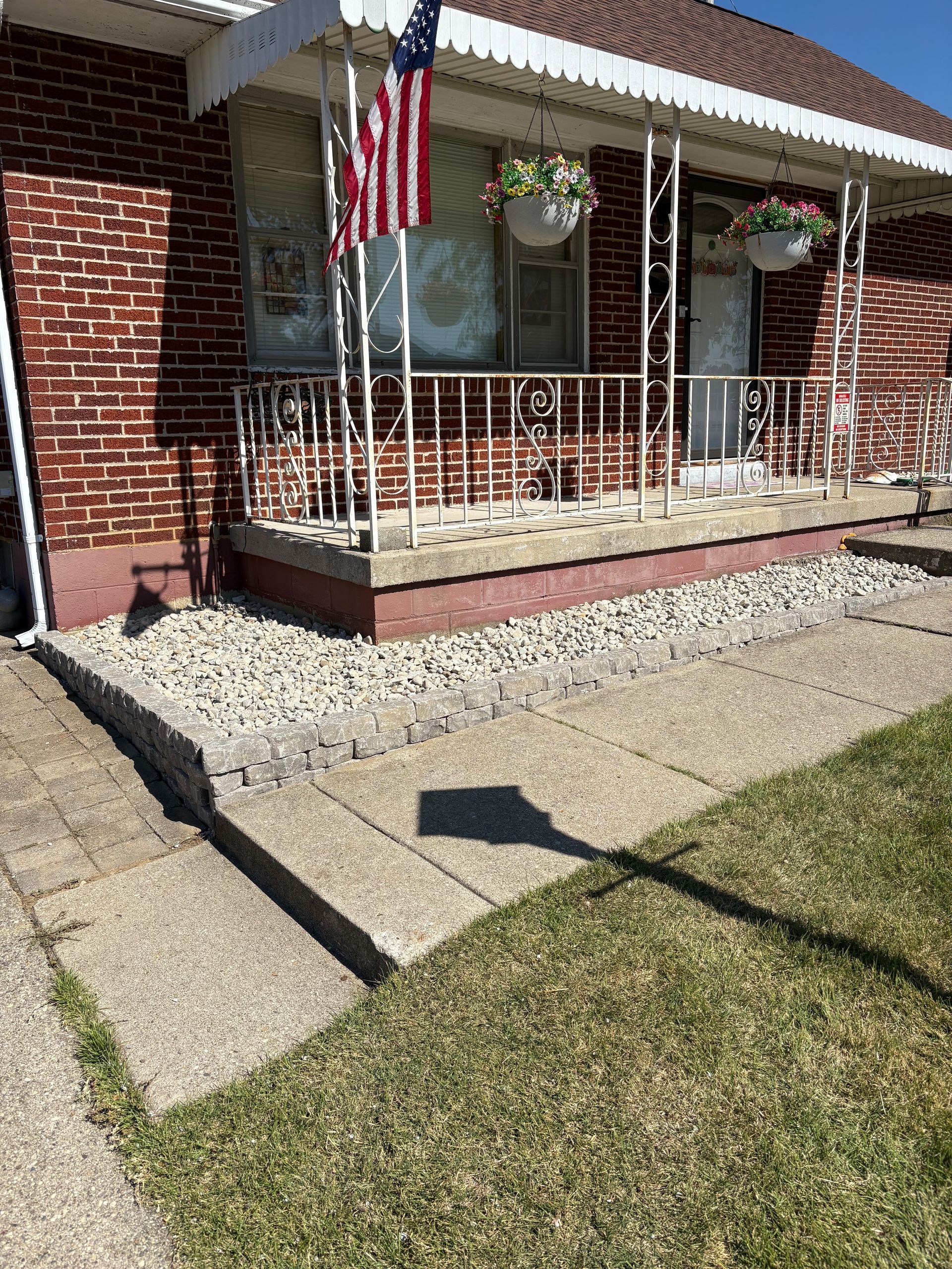 Brick house with a small porch and American flag, bordered by stones and a concrete sidewalk.
