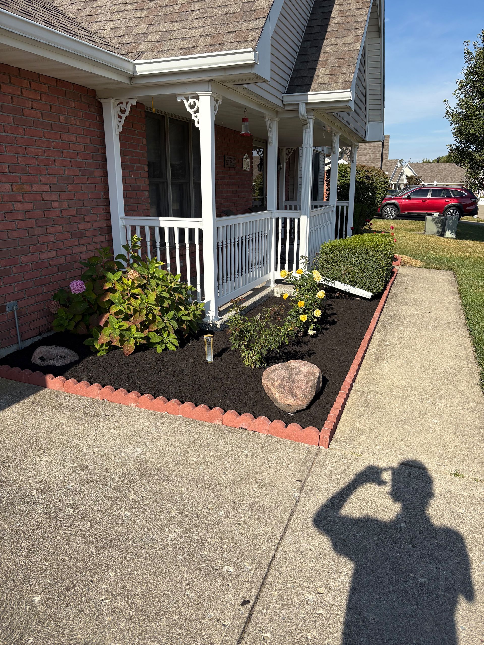 A house with a porch, surrounded by a brick border and landscaped garden, a person’s shadow is visible on the sidewalk.