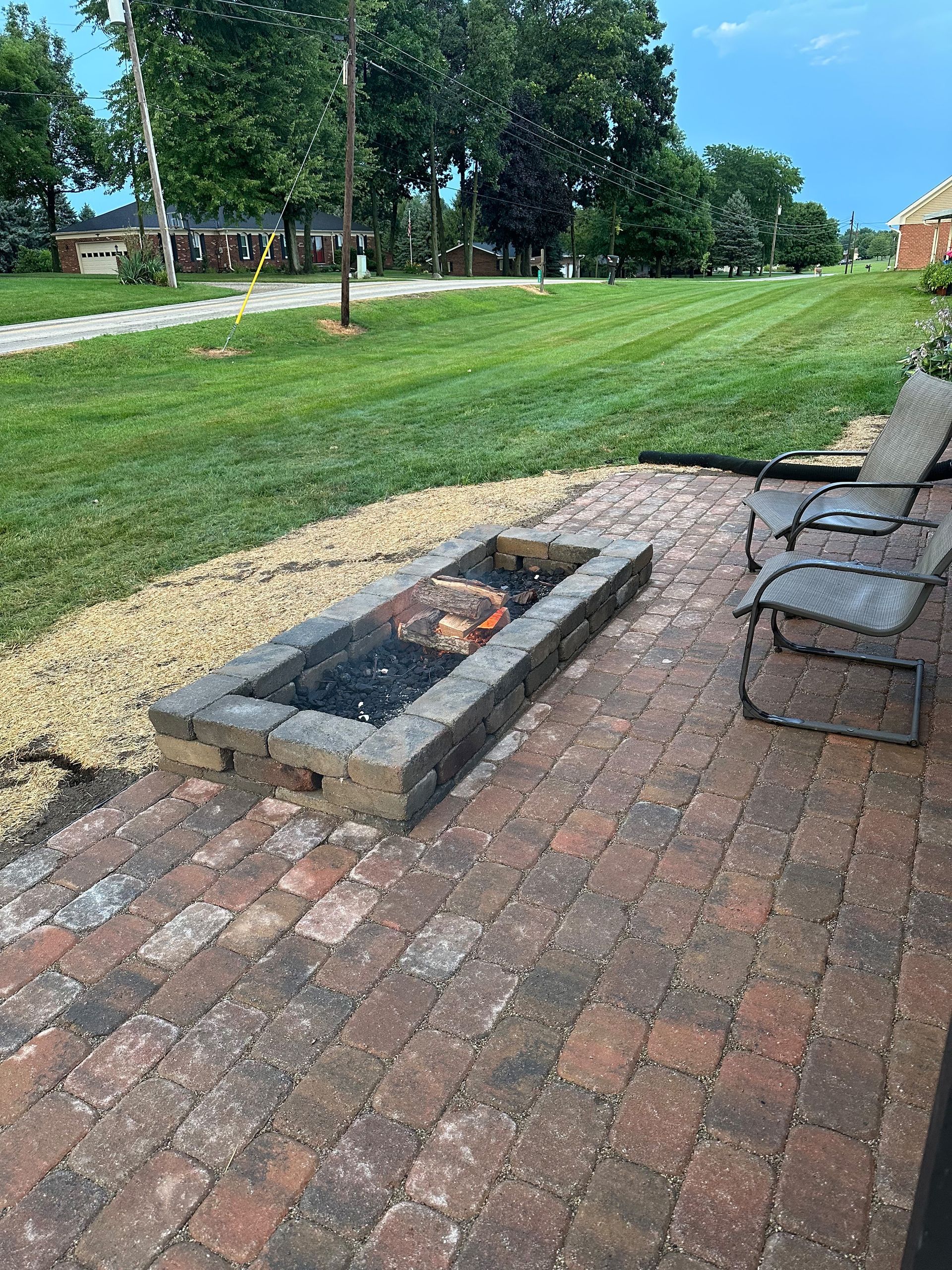 Brick patio with a rectangular fire pit, two chairs, and a grassy lawn in the background.