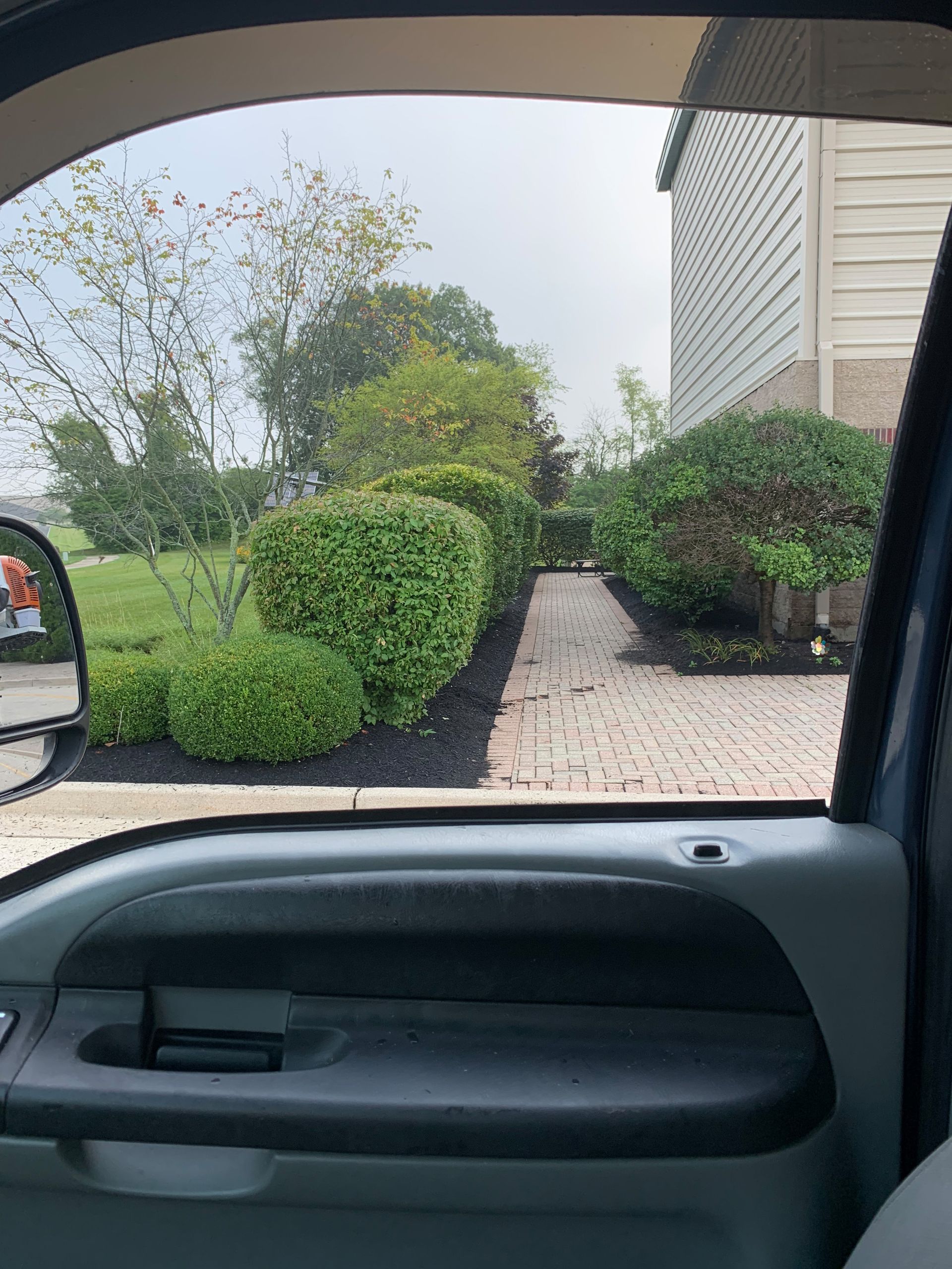 View from a car window of a manicured landscape with bushes, a brick path, and a house.