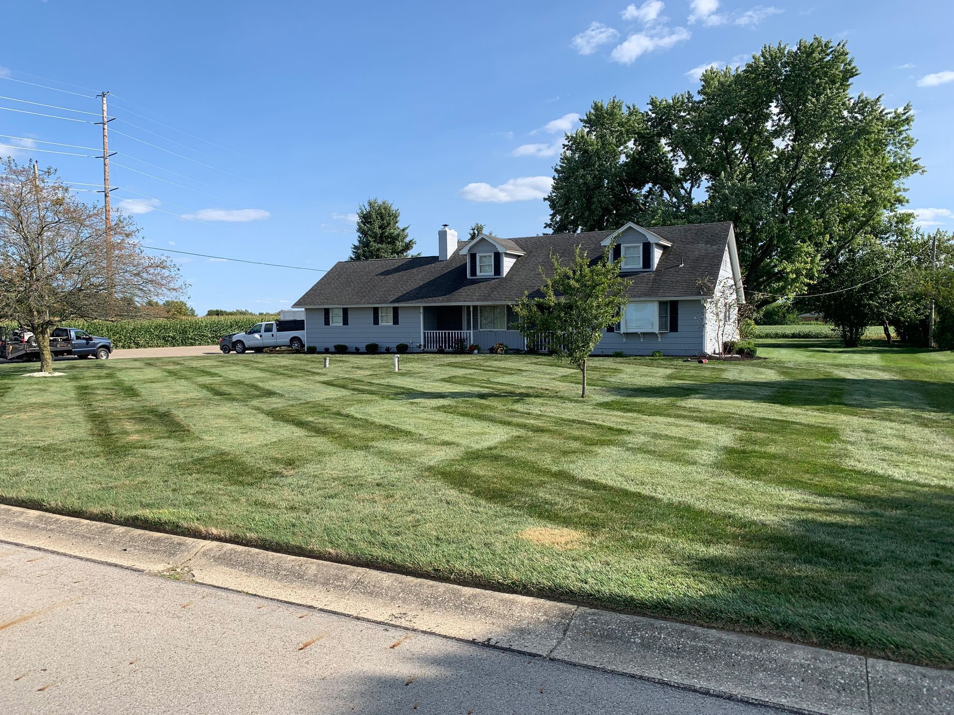 Gray house with a manicured lawn, blue sky, and a row of cornfields.