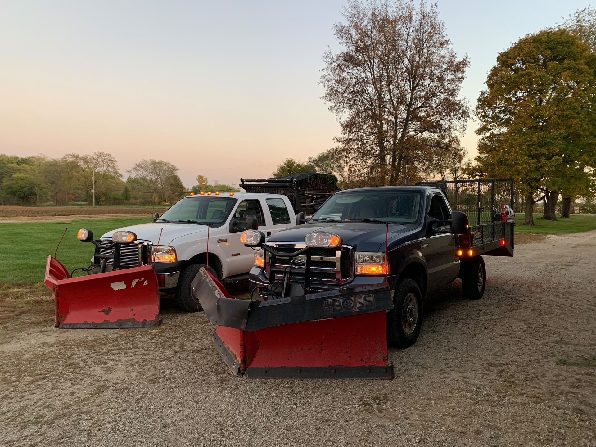 Two pickup trucks with snowplows parked on gravel, ready for winter.