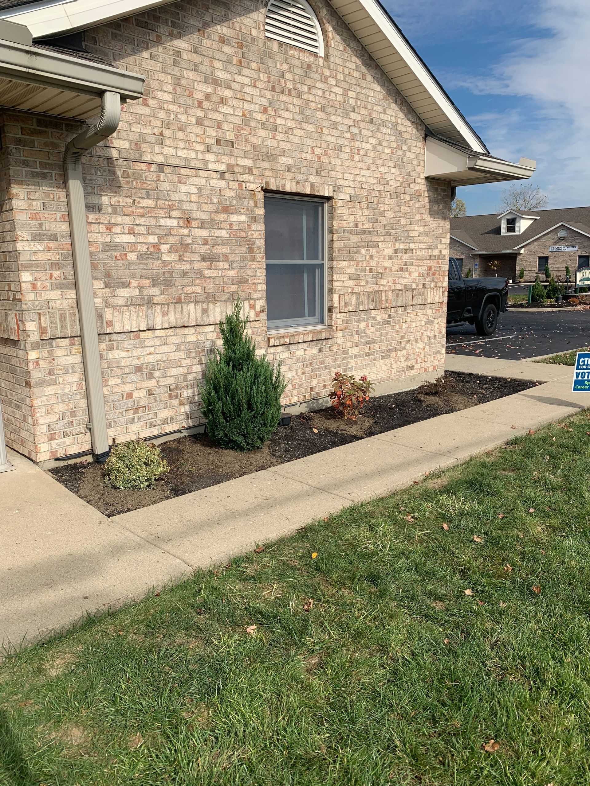 Brick building with a small evergreen bush and flower bed along a concrete sidewalk and grass.