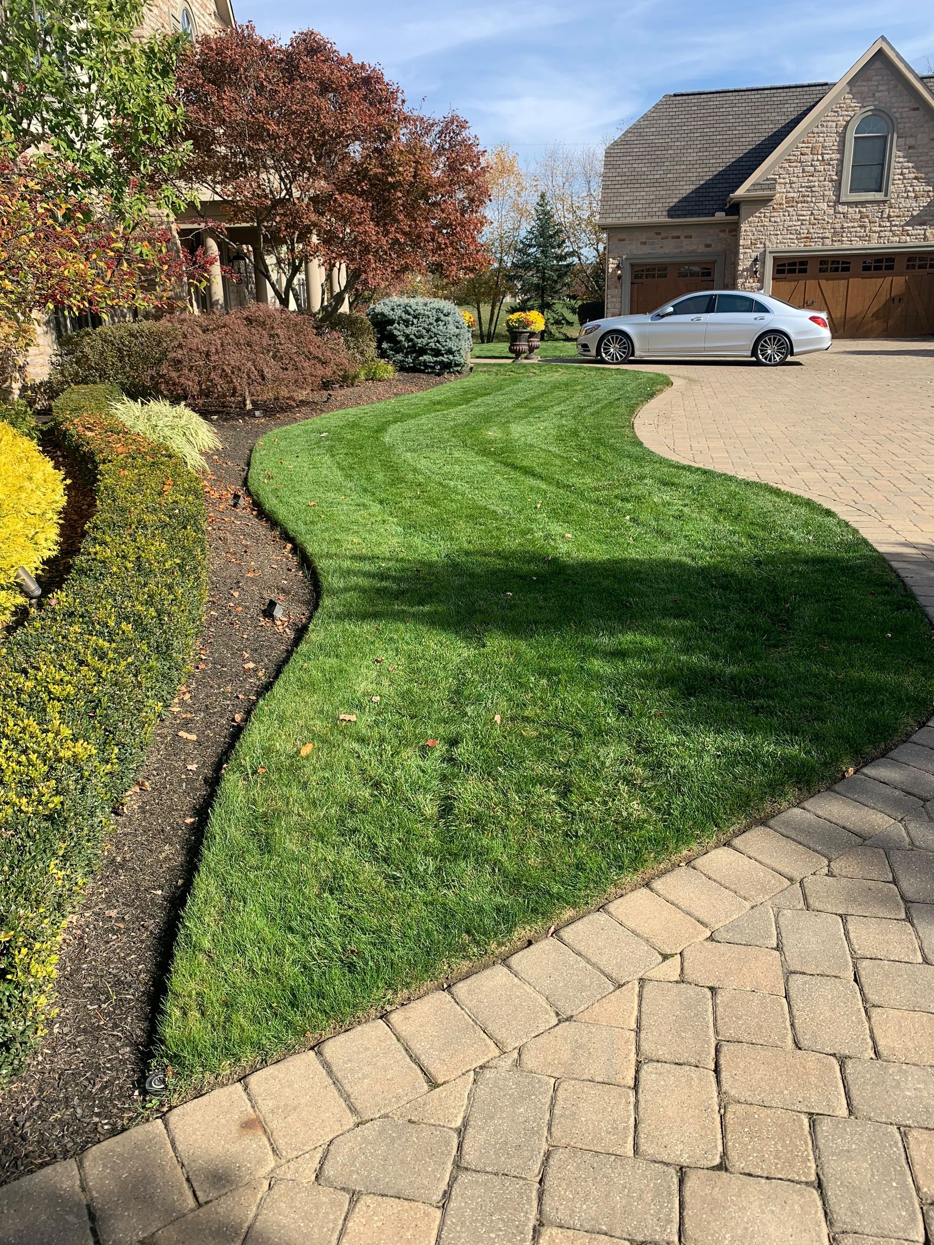 Lush green lawn curves beside brick walkway and flowerbeds. A white car sits in front of a stone house.