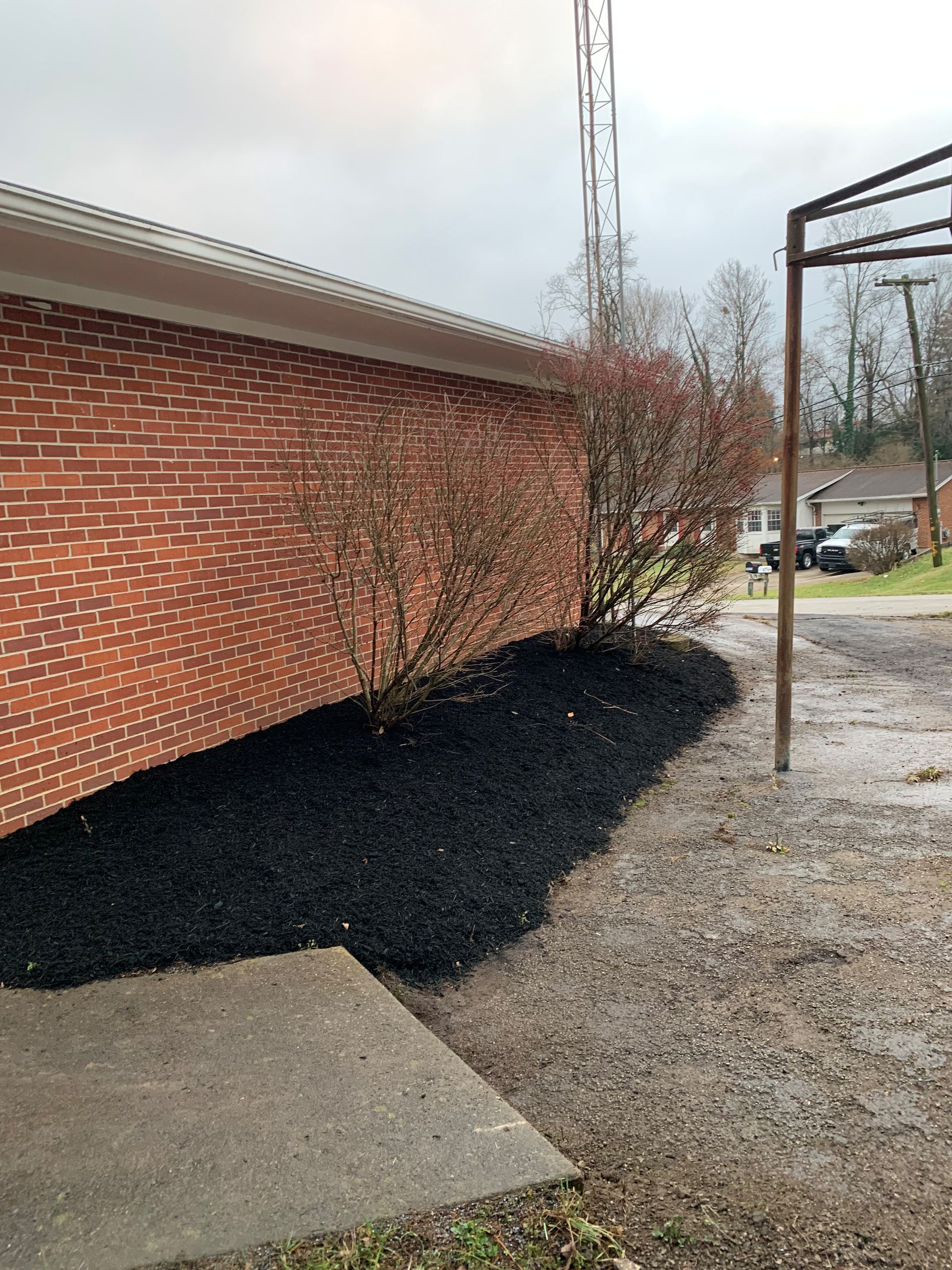 Brick building with a dark mulch bed and bare bushes along the wall, next to a gravel area.