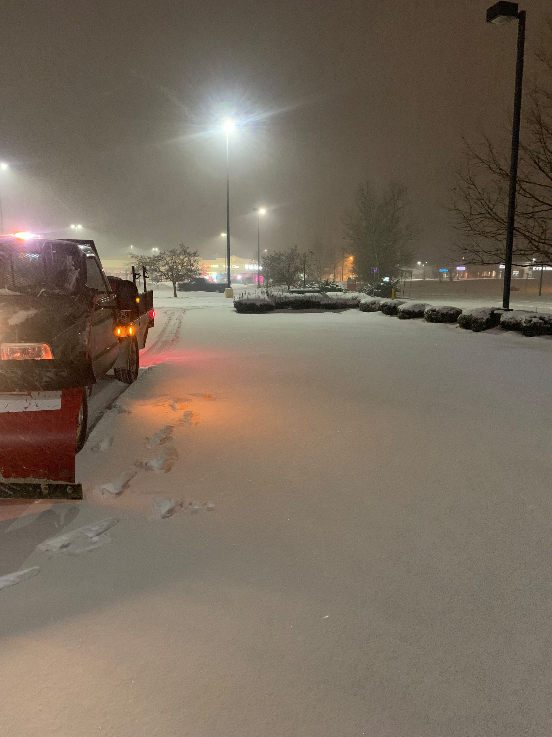 Snowplow clearing a snow-covered parking lot at night; streetlights illuminate the scene.