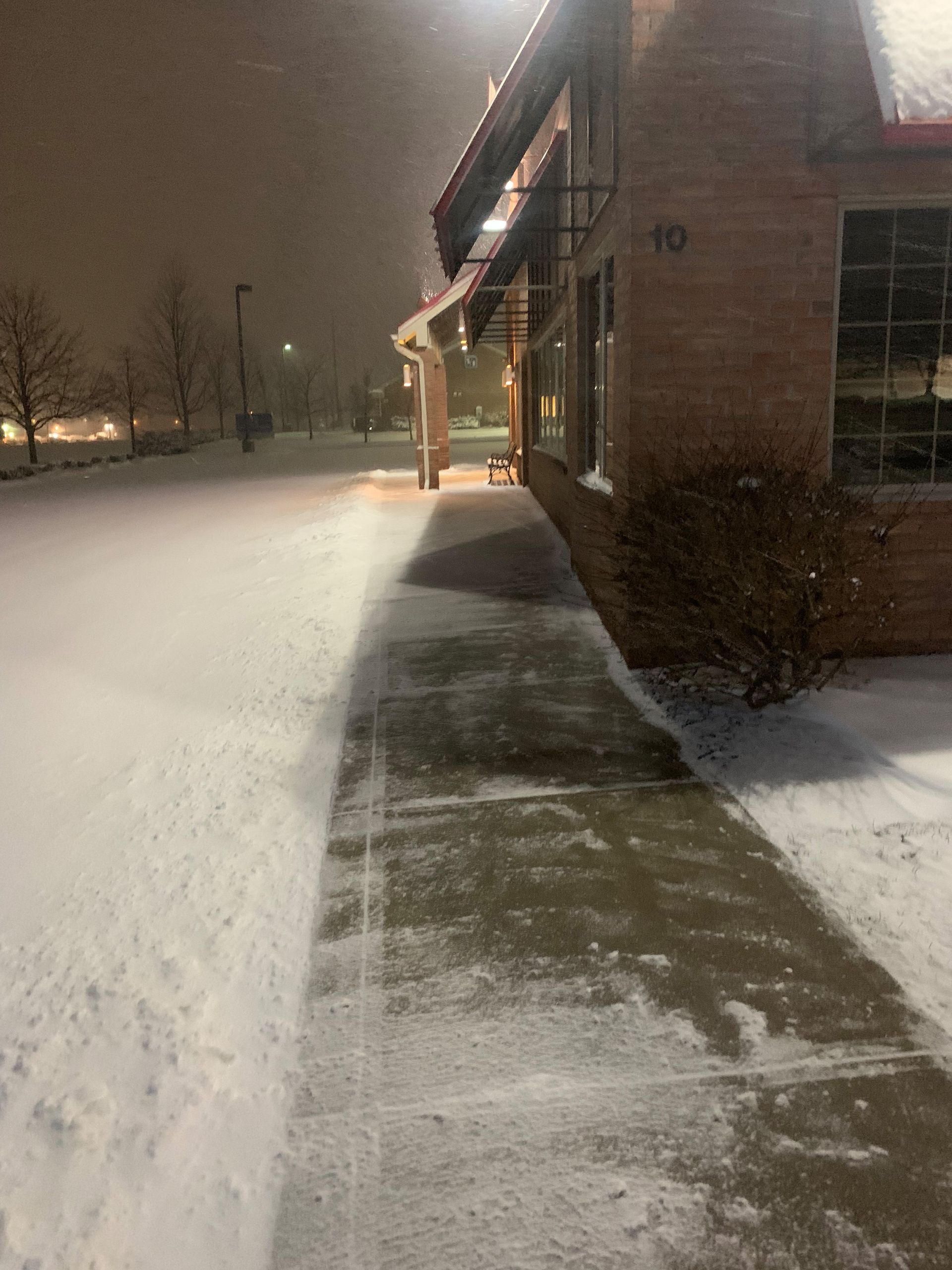 Snowy sidewalk next to a brick building at night. Snow covers ground and obscures trees in the distance.