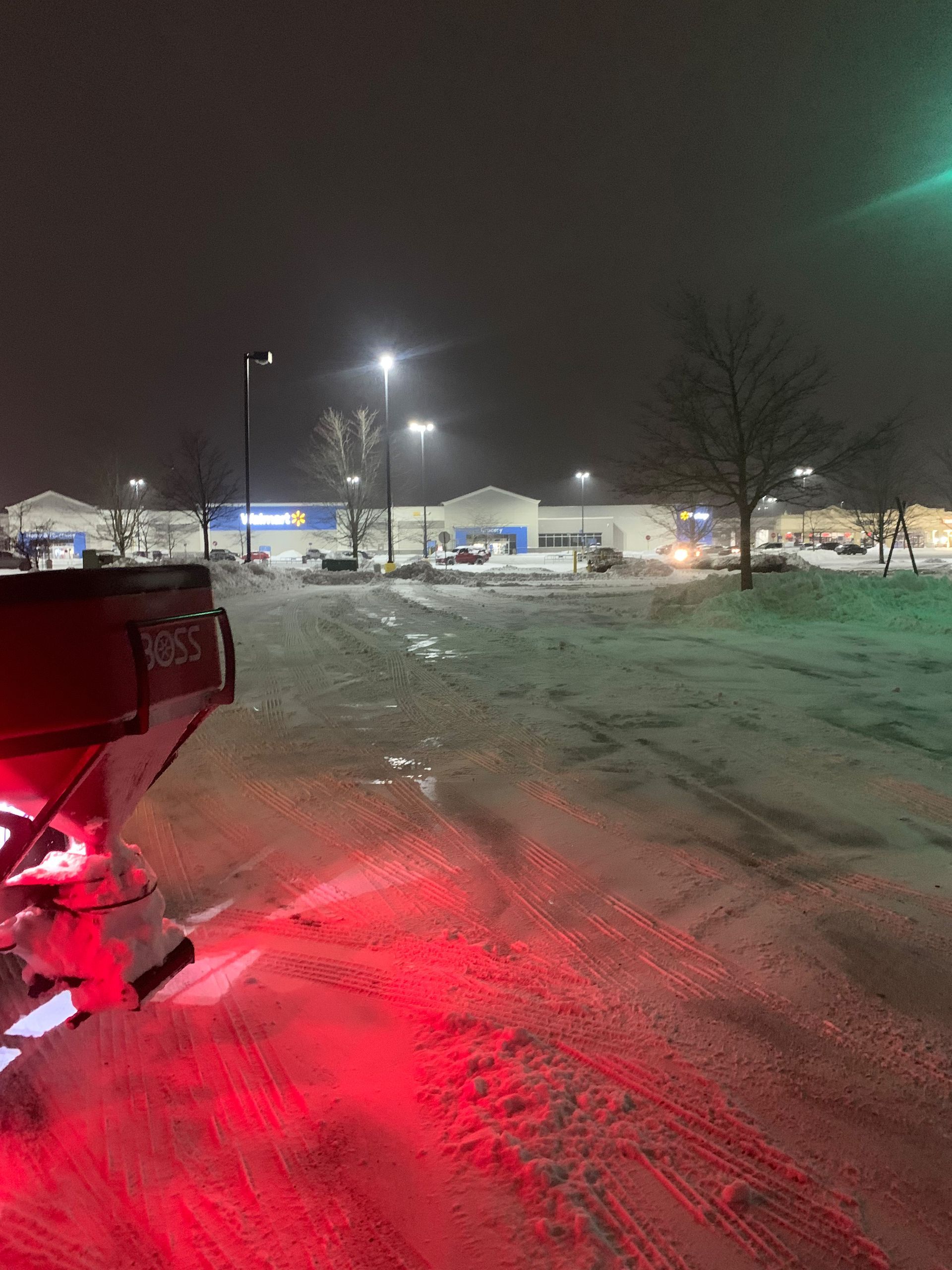 Snowplow clearing a snowy parking lot in front of a Walmart store at night; red lights glow.