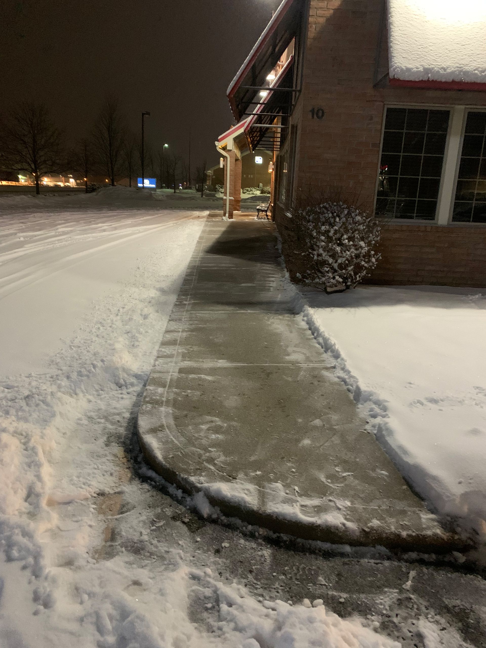 Snowy sidewalk next to a building at night. A small shrub sits near the building's entrance.