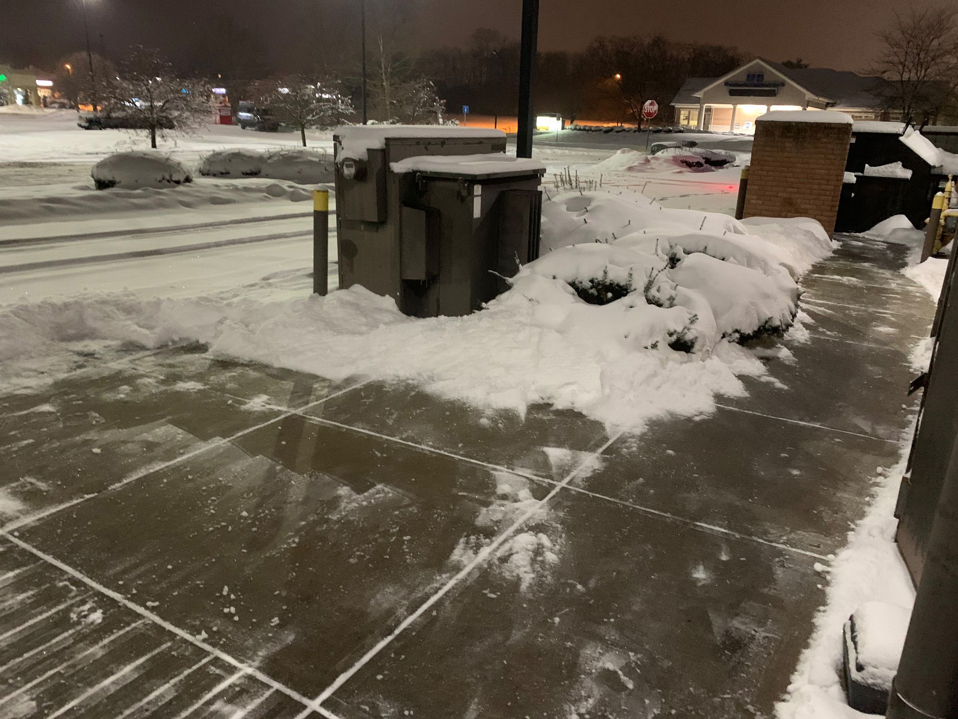 Snow-covered sidewalk and parking lot at night; an electrical box and bushes are covered in snow.