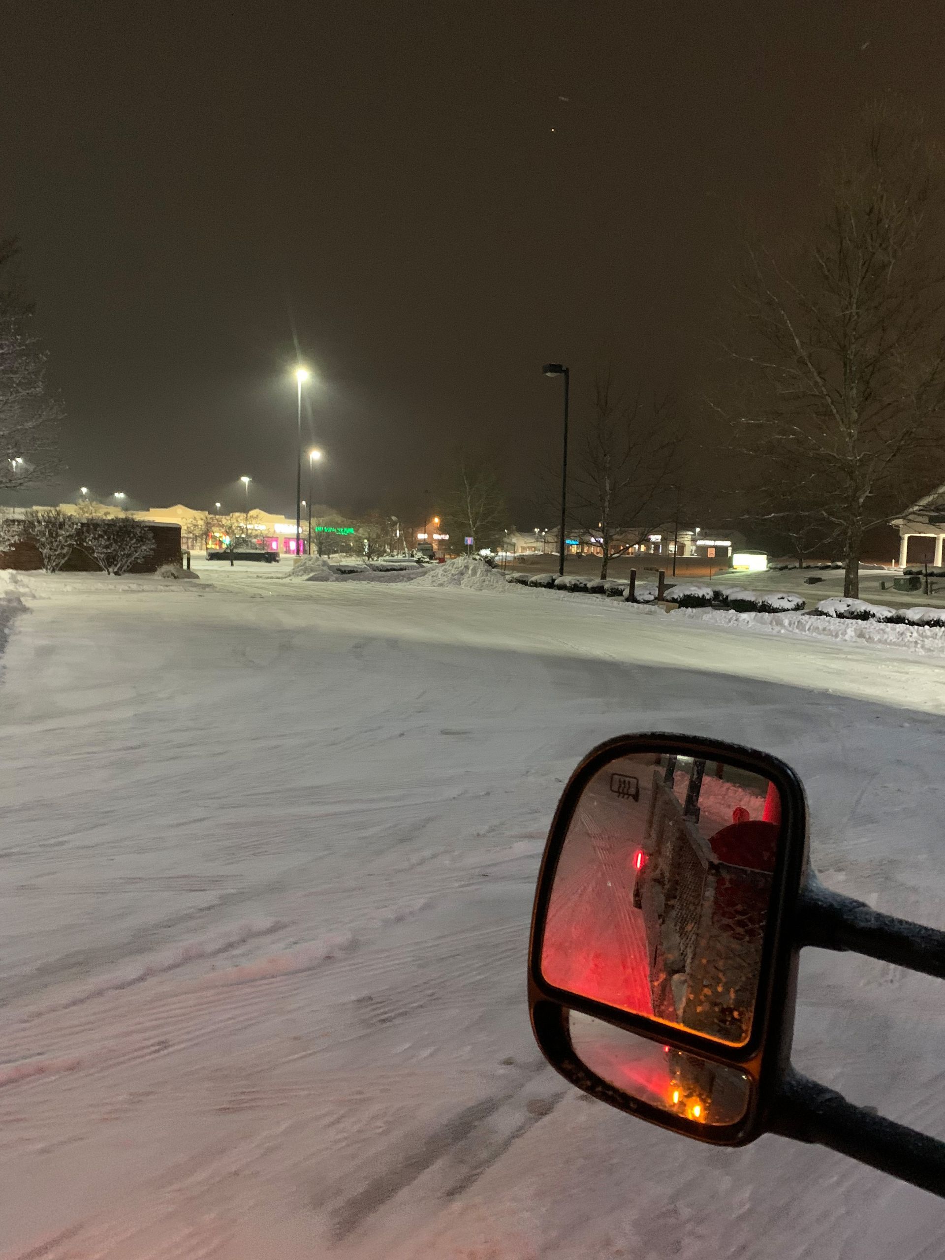 Snow-covered road at night, seen from a vehicle. Snow flurries, streetlights, and building lights in the distance.