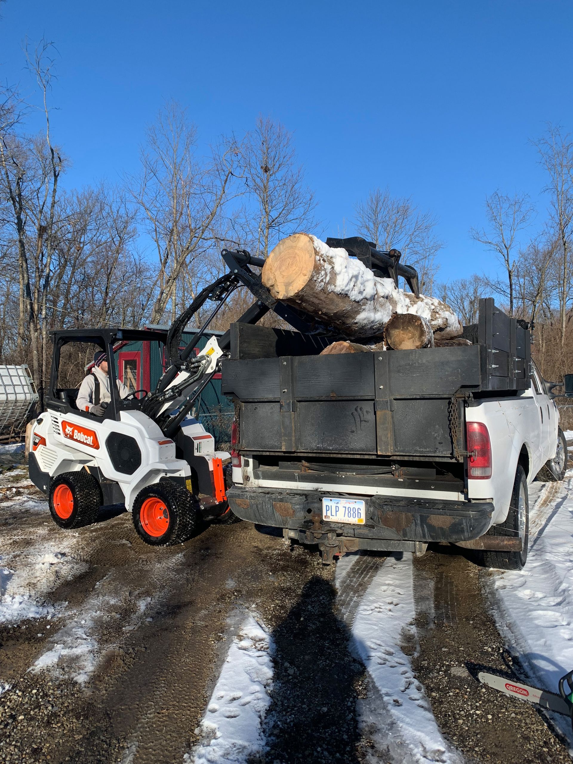 A white Bobcat skid steer loads a large log into a white pickup truck on a snowy ground.