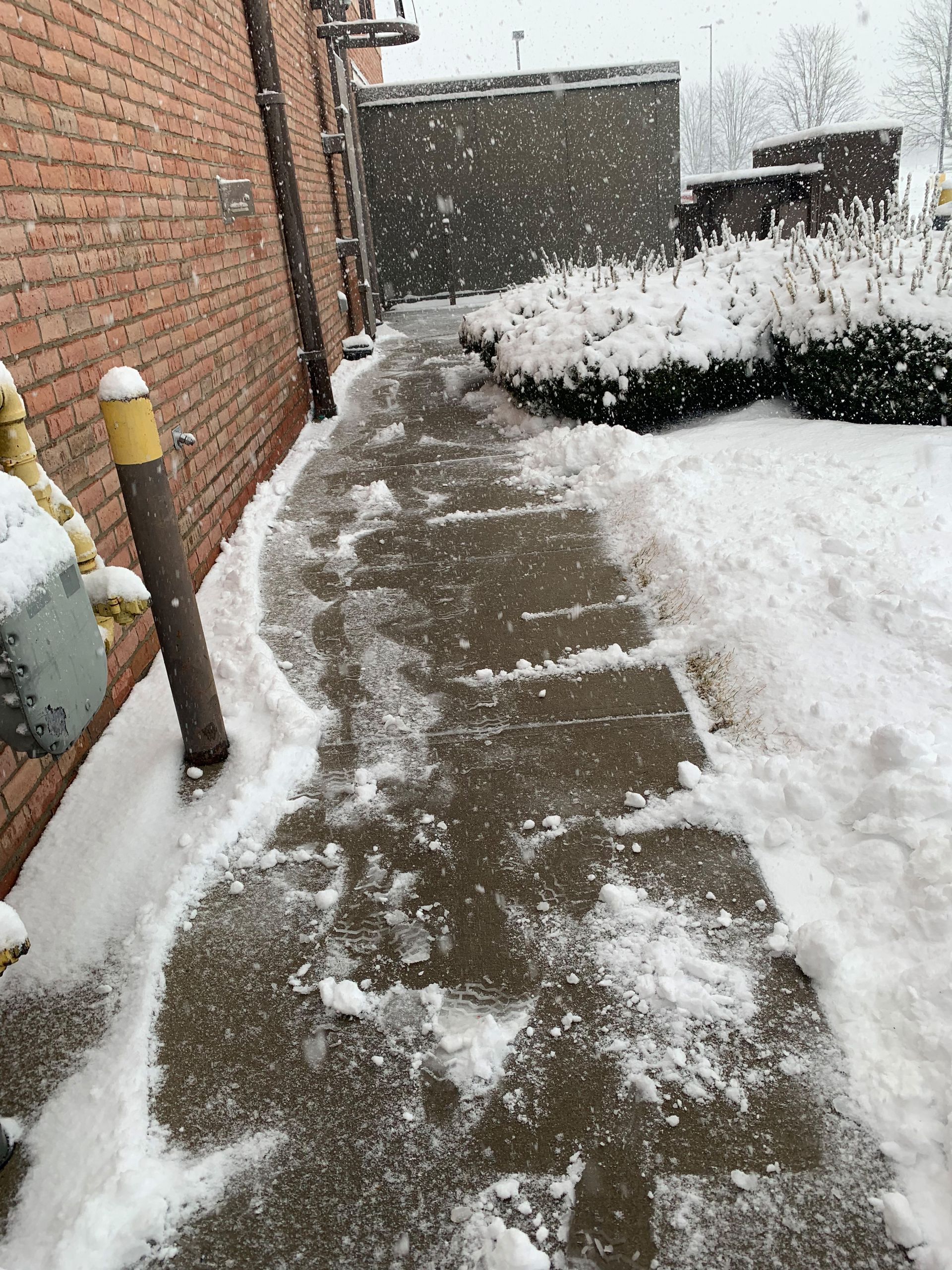 Snowy sidewalk with footprints, next to a brick building. Snow falling.