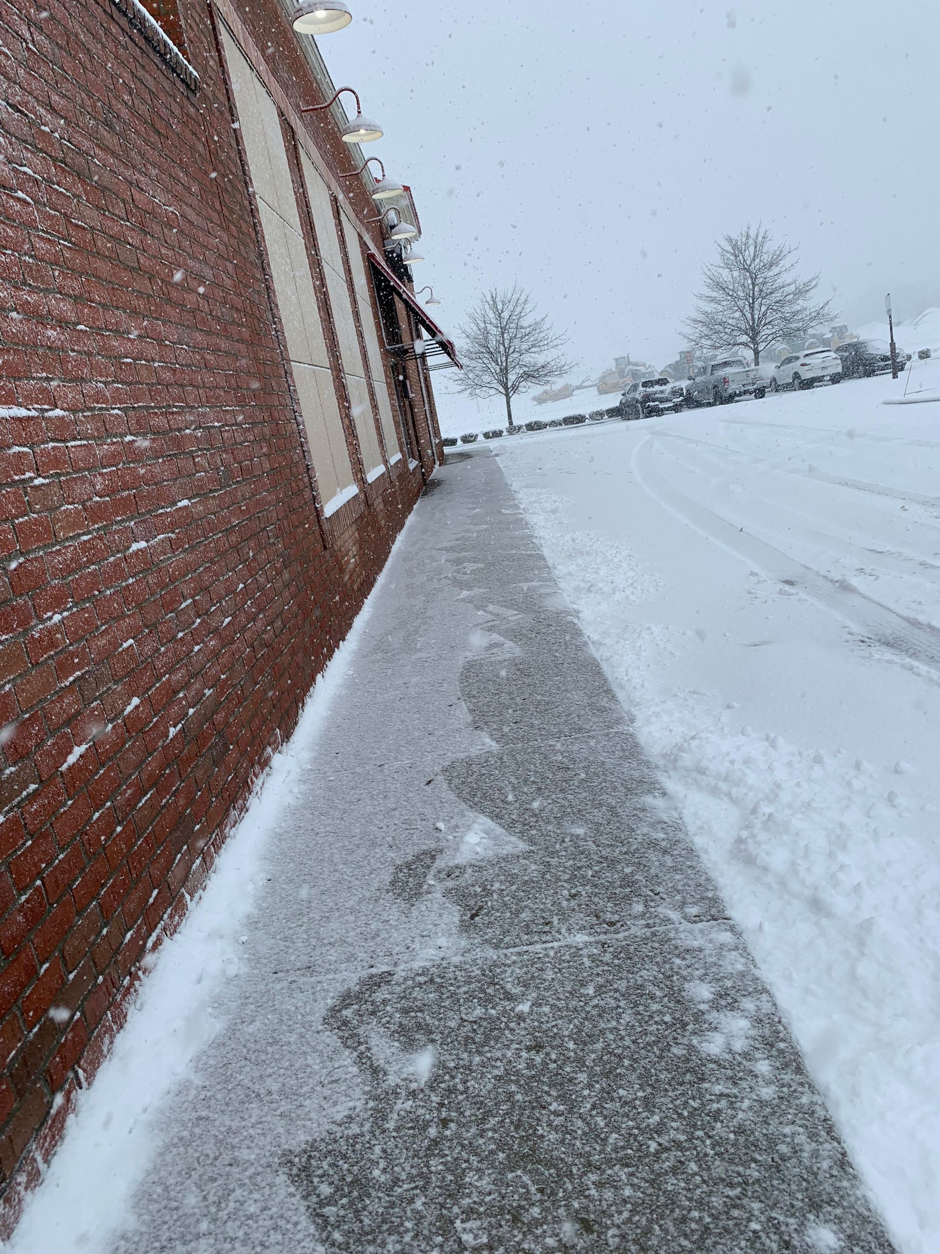 Snowy sidewalk next to a brick building. Trees and a road in the background.