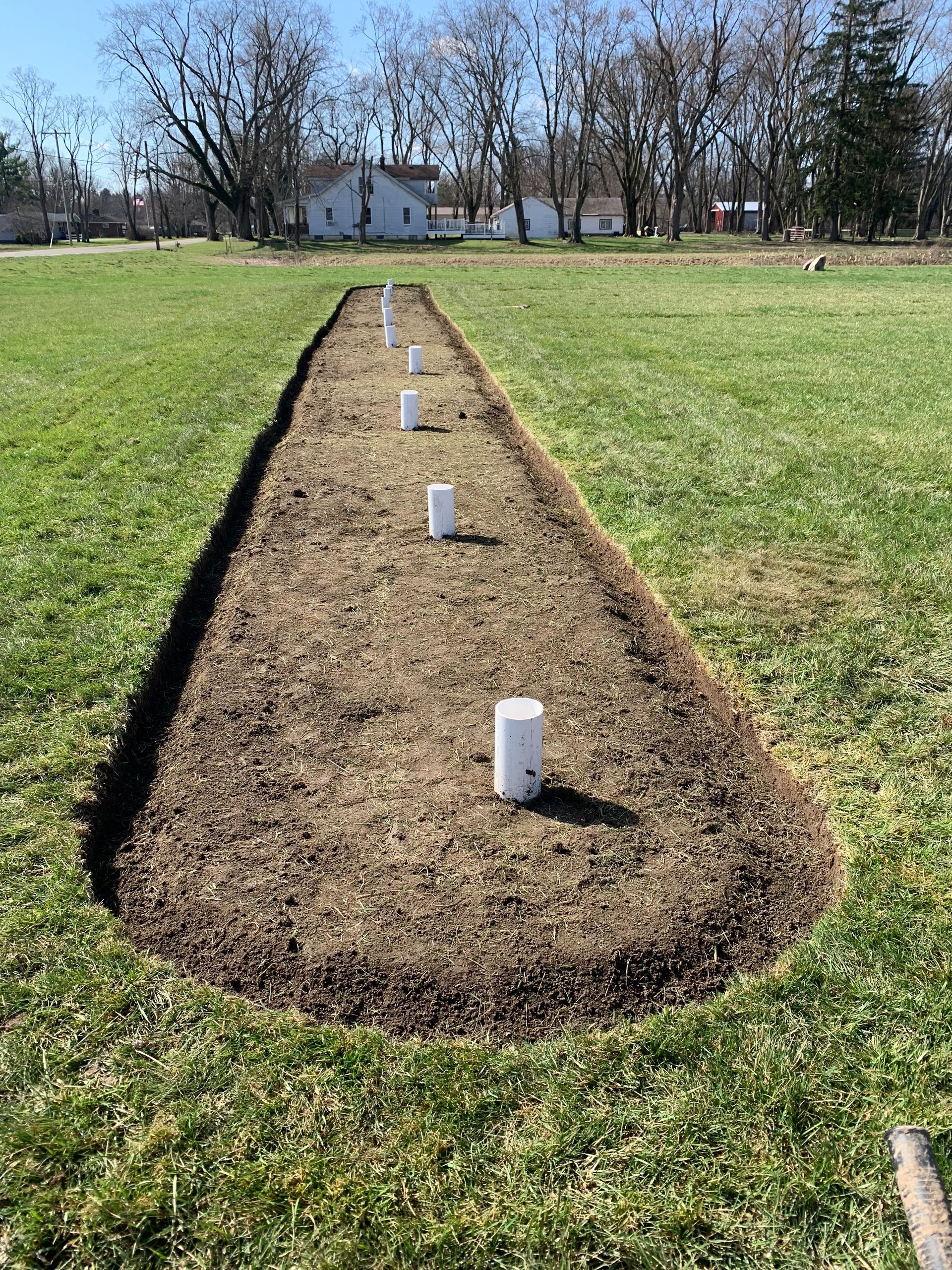 A long, mulched garden bed with white pipes, in a grassy field, houses visible buildings in the background.